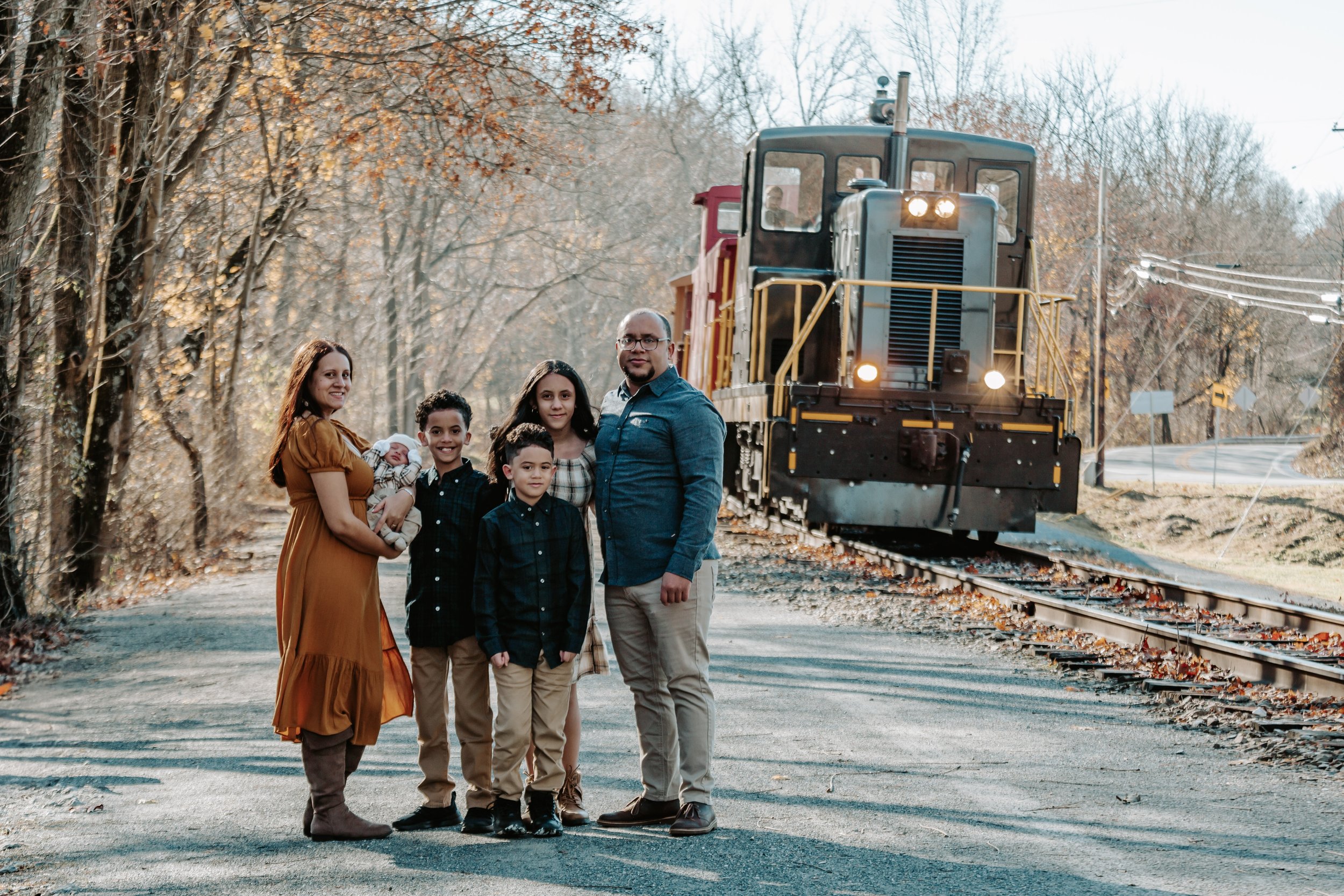 A family of six standing on a road beside a train track with a train behind them, surrounded by autumn trees with leaves fallen on the ground.