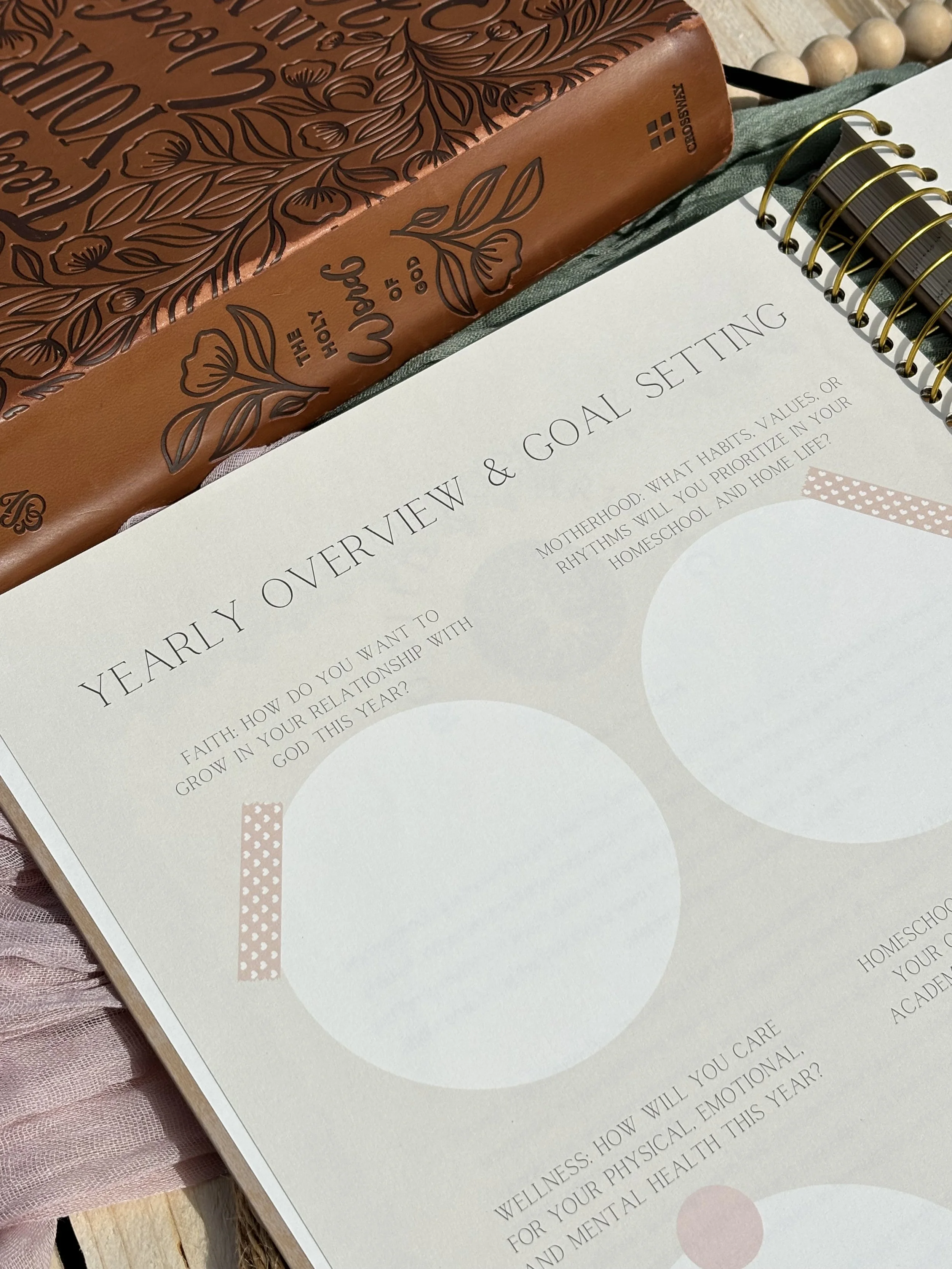 A notebook or planner with a brown leather cover, decorated with embossed floral patterns and the phrase "The Holy Word of God." Next to it is an open worksheet titled "Early Overview & Goal Setting" with questions about health, wellness, and relationship goals for the year, accompanied by a wooden beaded bracelet and a spiral-bound planner or notebook with gold rings. A pink tulle fabric is partially visible underneath on a wooden surface.