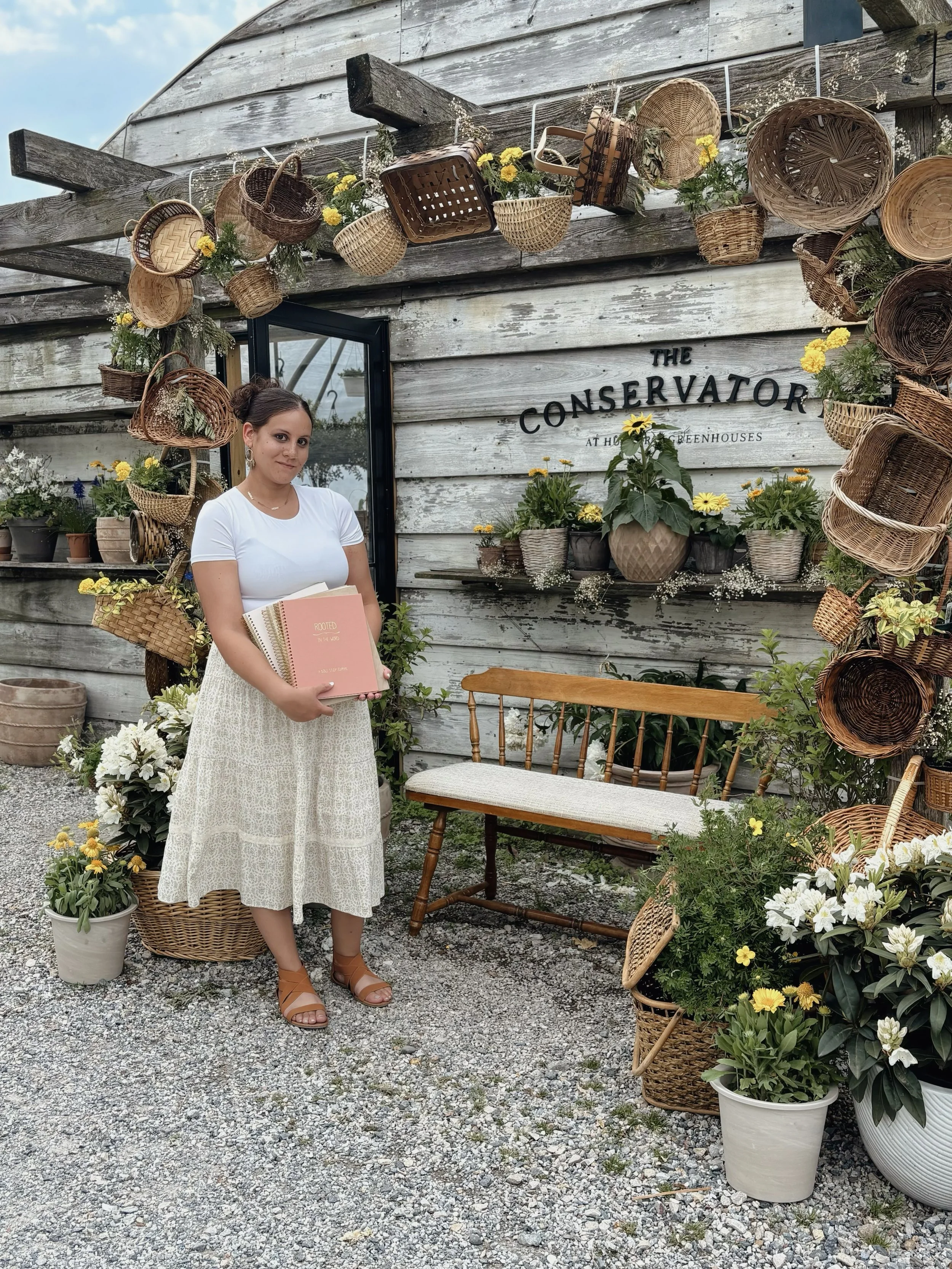 A young woman standing in front of a wooden wall decorated with hanging baskets and flowers outside a greenhouse. She is holding a notebook and smiling slightly, wearing a white t-shirt and a long cream-colored skirt. There are potted flowers and plants around her, and a wooden bench nearby.