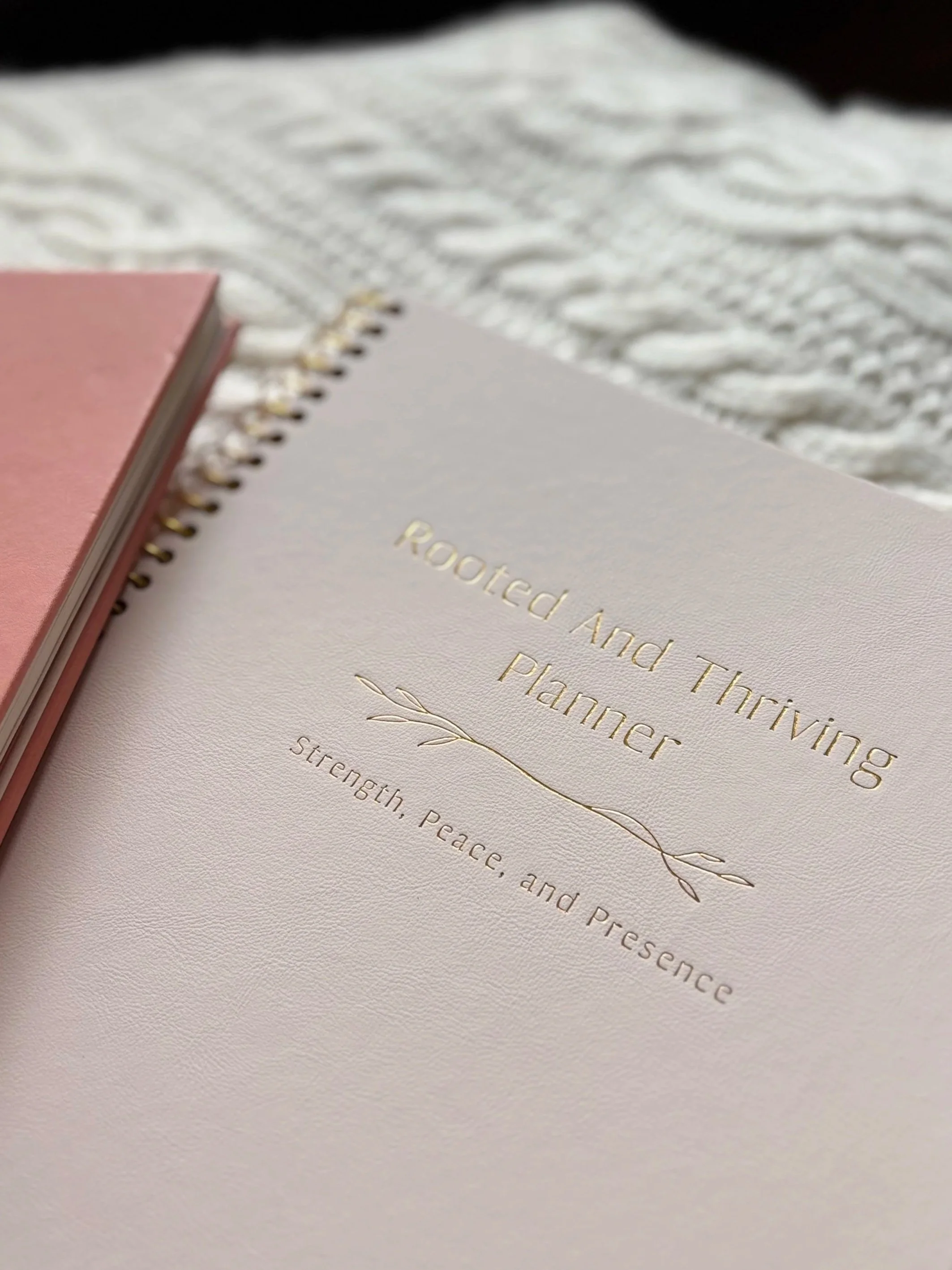Close-up of three notebooks with pink, white, and patterned covers on a textured white surface, with gold embossed text on the white notebook about strength, peace, and presence.