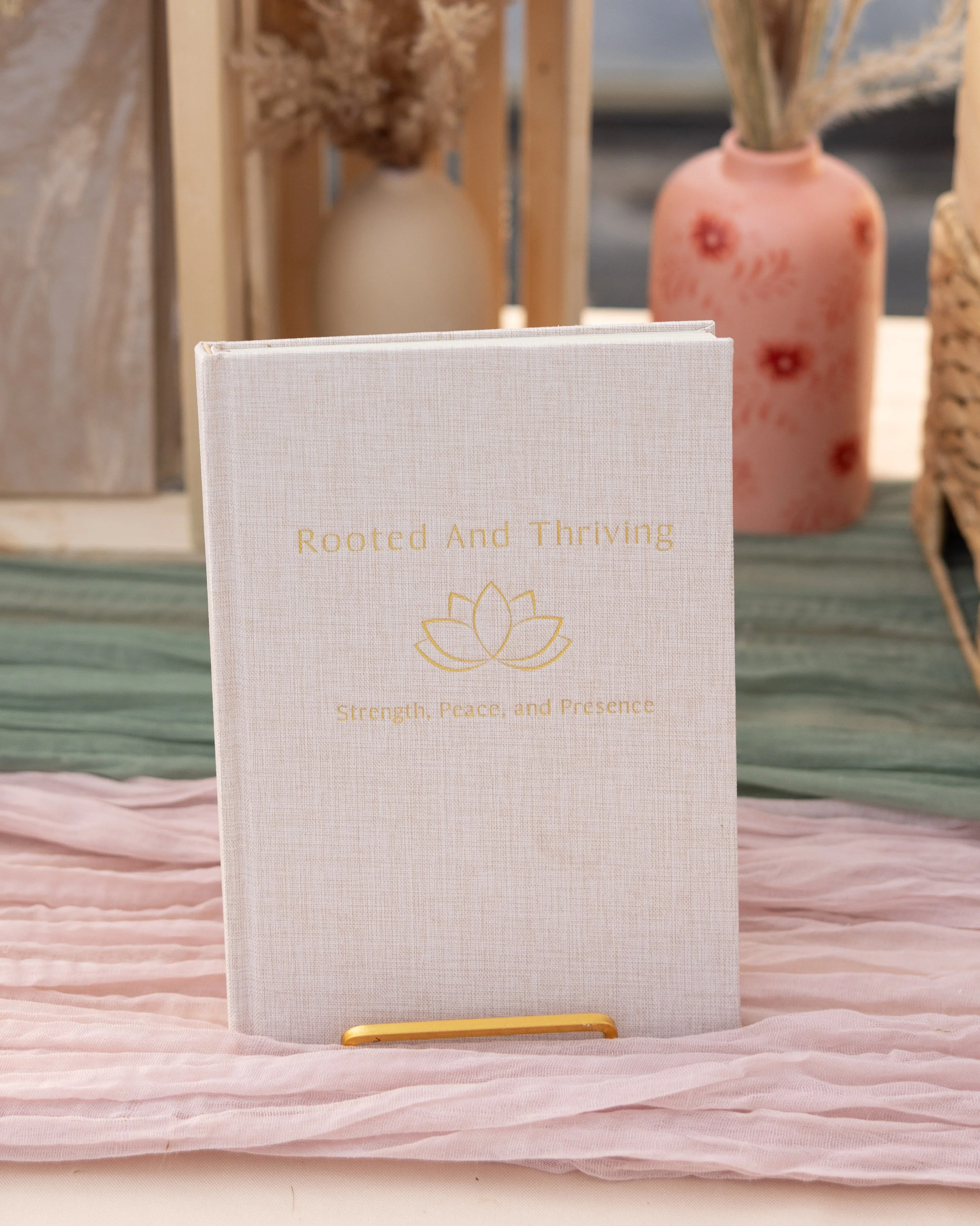 A white journal titled 'Rooted and Thriving' with a subtitle 'Strength, Peace, and Presence' and a lotus flower illustration, displayed on a pink fabric surface, with decorative vases and flowers in the background.