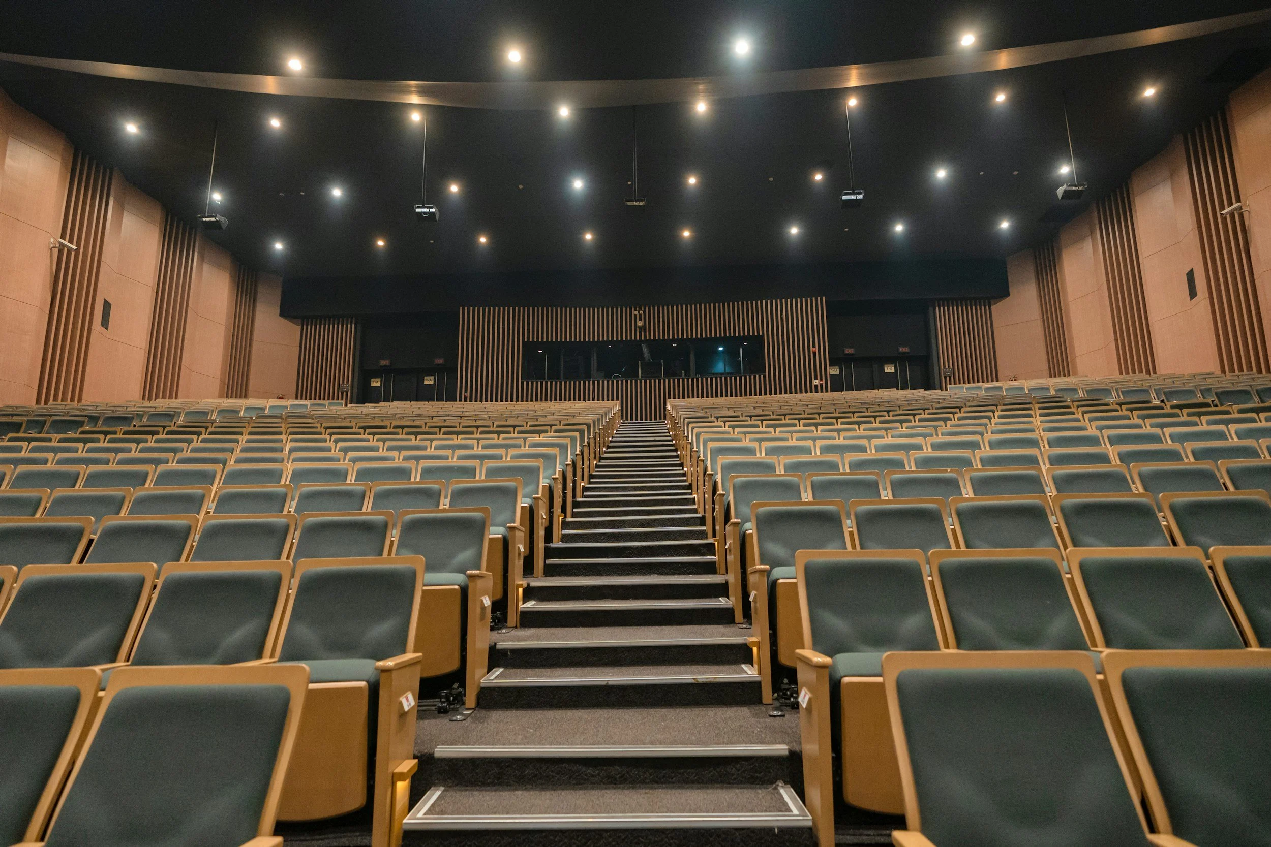 Empty auditorium with rows of green seats, a central staircase, and a black stage with a large window behind it, illuminated by ceiling lights.