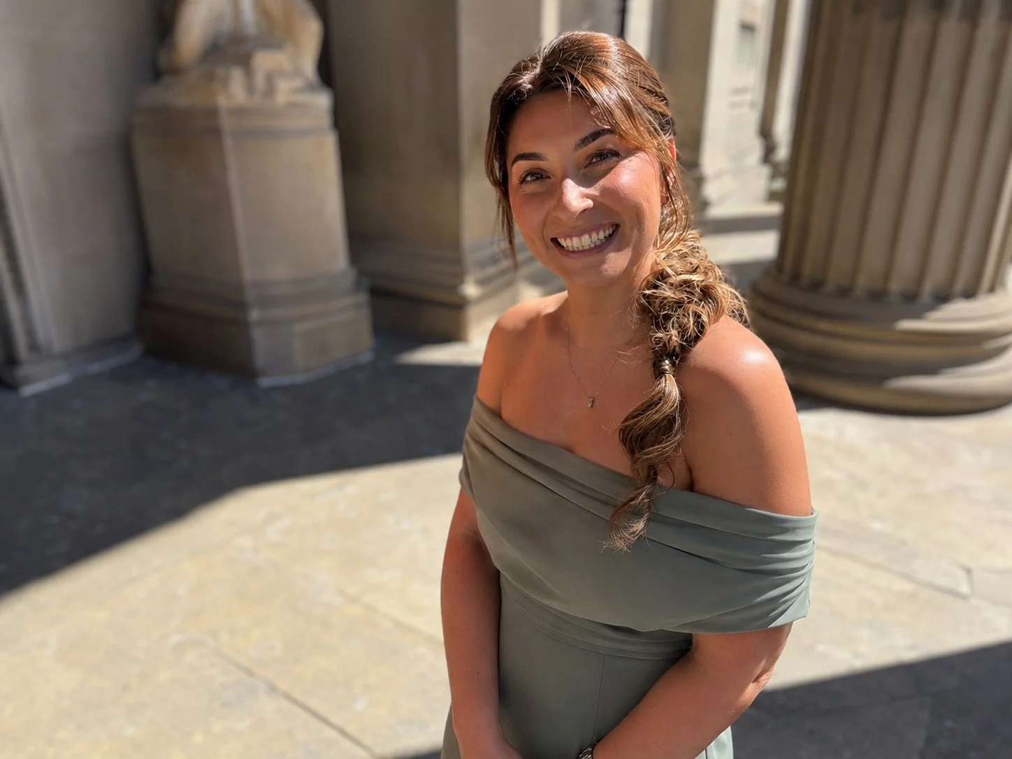 A woman with light brown hair styled in a side braid, wearing a gray off-shoulder dress, smiling at the camera, standing outside against a background of classical architectural columns.