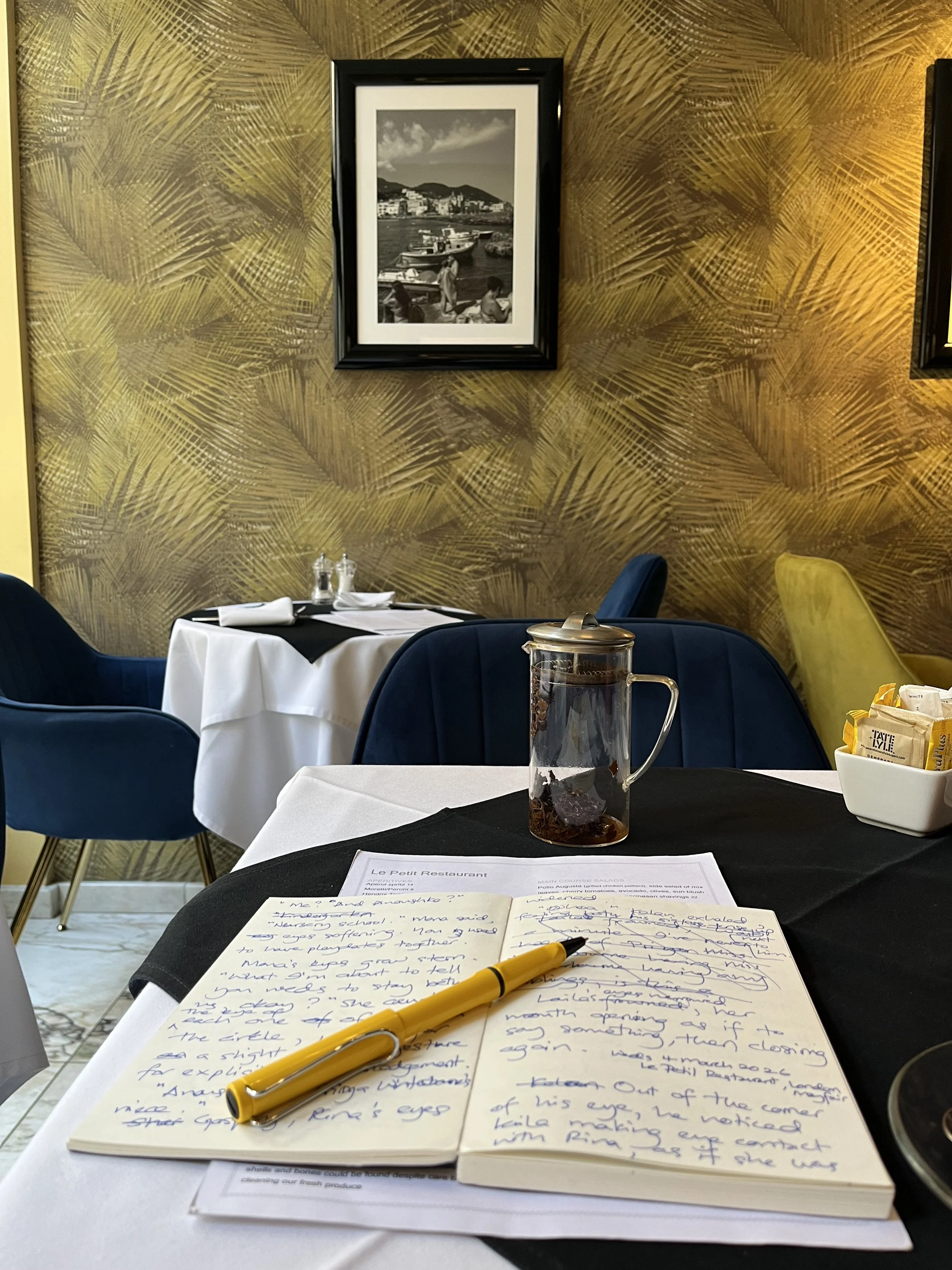 A table with a yellow pen and handwritten notes, a glass container with a dark liquid, and a white bowl with sugar packets at a restaurant. In the background, a framed black-and-white photo of boats at a harbor hangs on a yellow palm leaf-patterned w