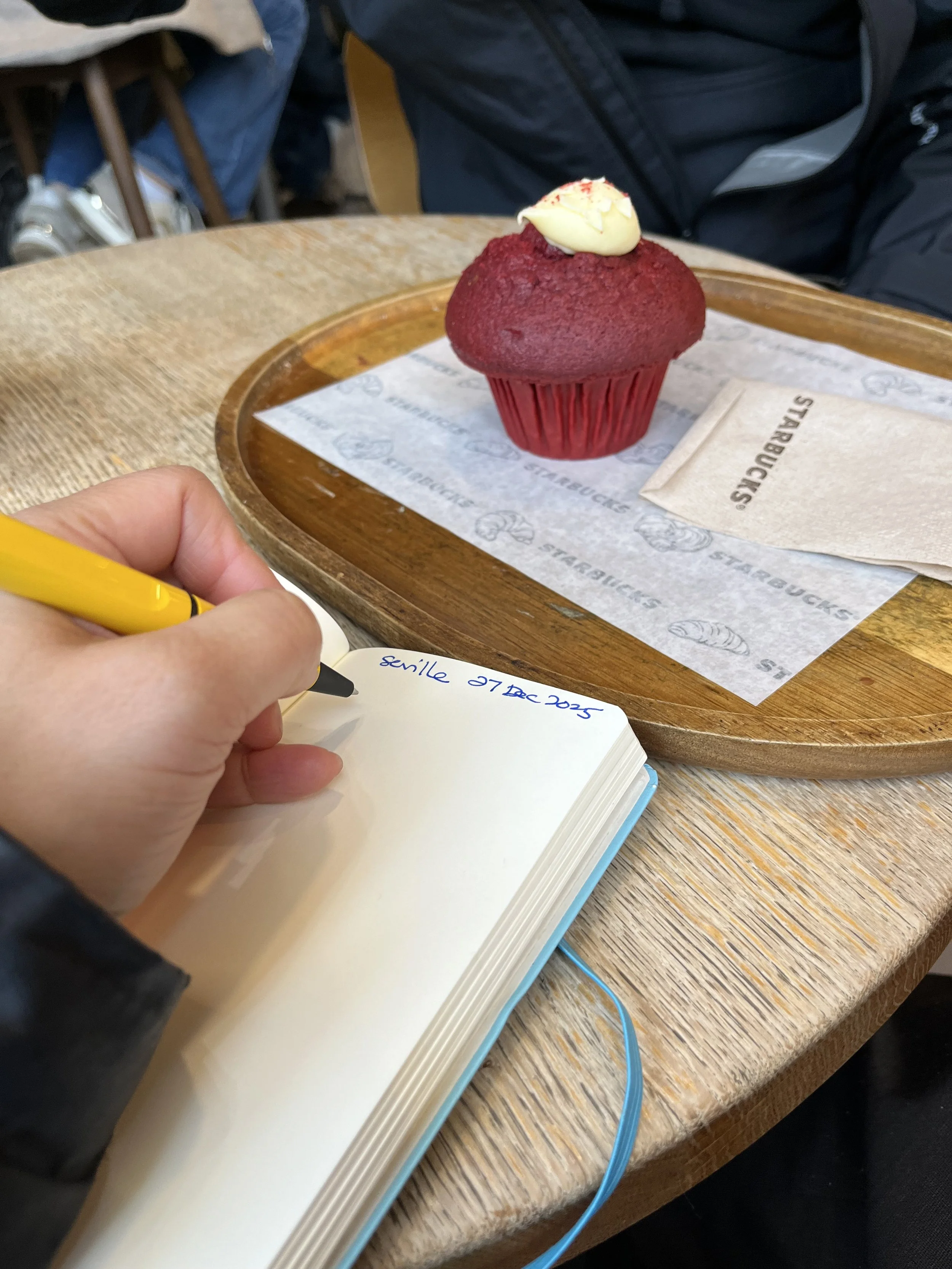 A red cupcake with a white dollop of frosting on top on a wooden tray, with a person writing in a notebook nearby.