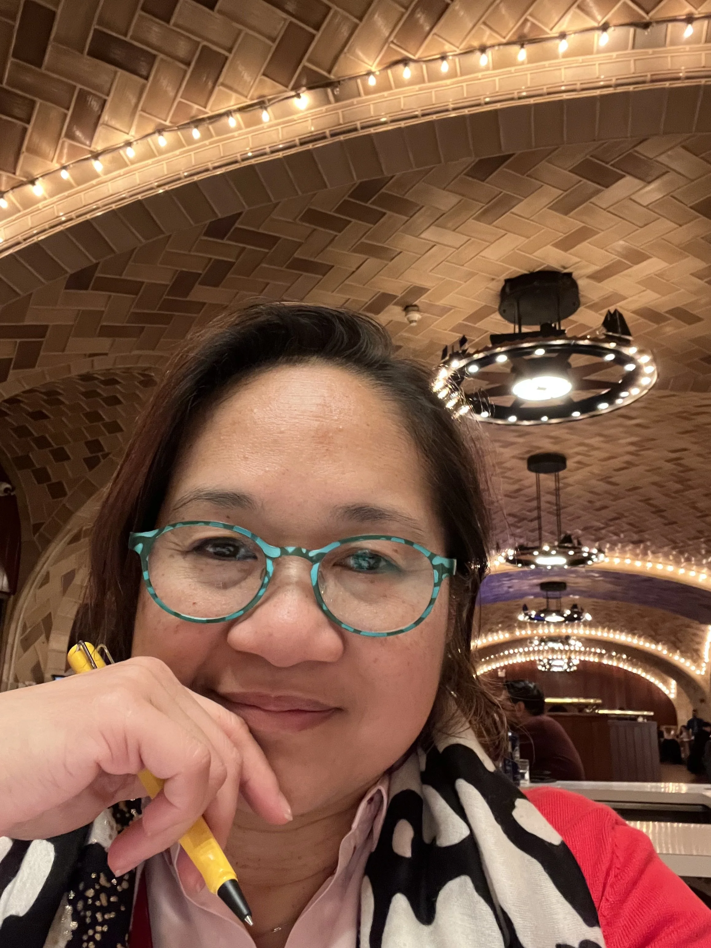 A woman with glasses and a patterned sweater smiling in a restaurant or cafe with a wooden arched ceiling and hanging lights.