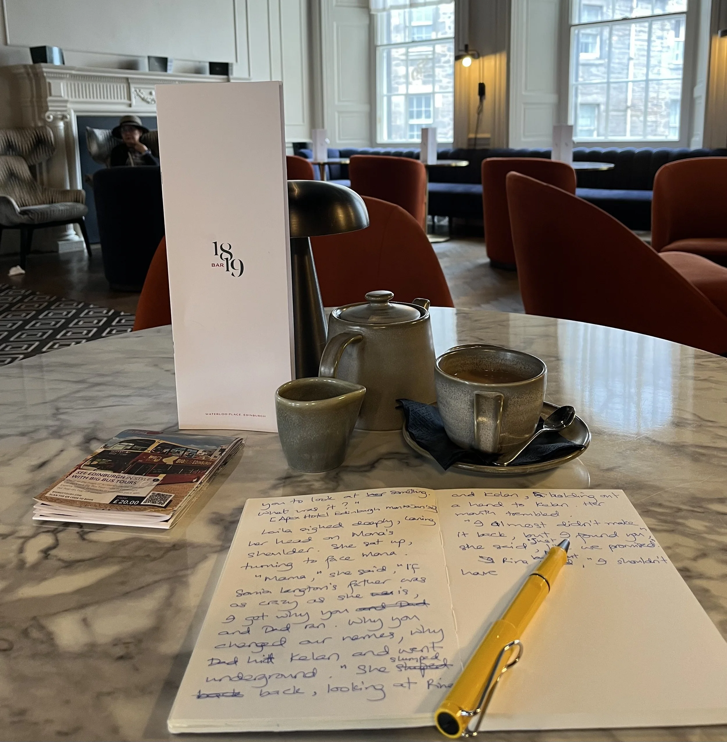A close-up of a marble table with handwritten notes on a white sheet of paper, a yellow pen, and a ceramic mug. In the background, a restaurant interior with large windows, orange chairs, wall lighting, and a person wearing a hat sitting in a cozy co