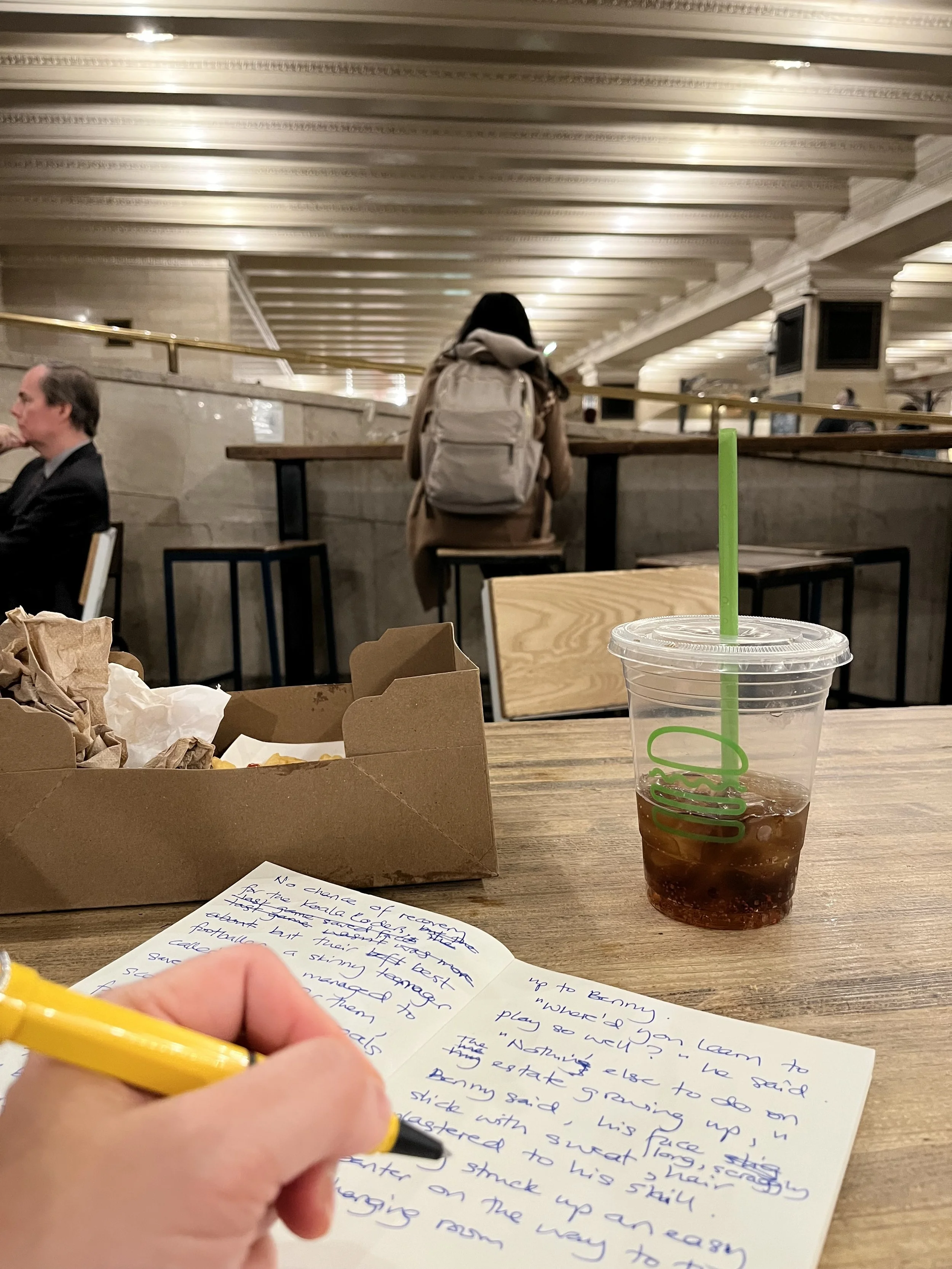 A picture taken from the person's perspective at a table in a busy indoor setting. In the foreground, a hand is writing on a notebook with a yellow pen. On the table, there is a clear plastic cup with a green straw and a dark-colored beverage. Also, 