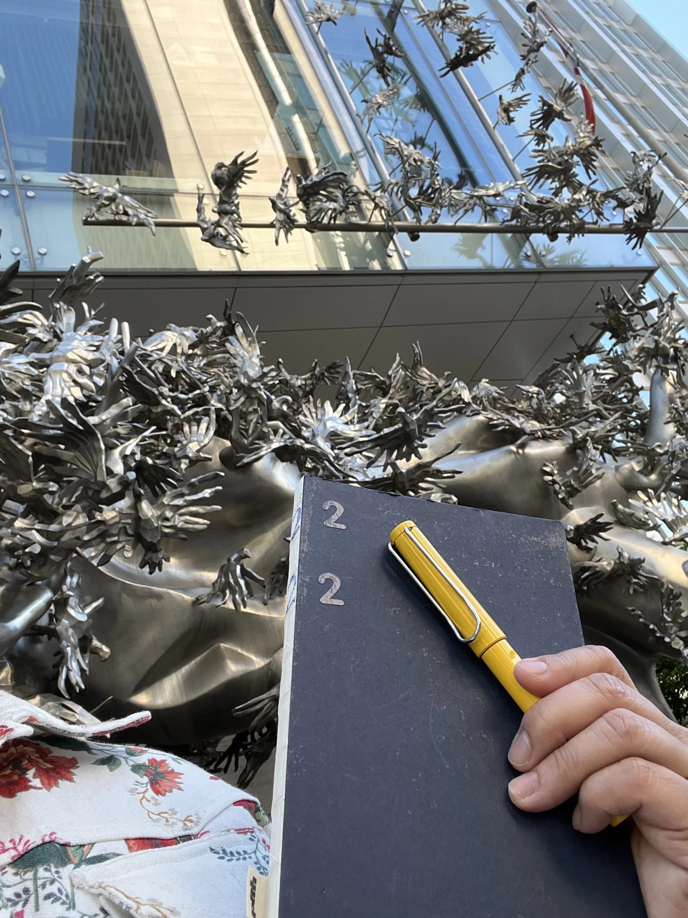 Close-up view of a hand holding a black sketchbook with a yellow pen on top, in front of an intricate metallic sculpture of numerous birds. The background shows a modern glass building with reflective surfaces.