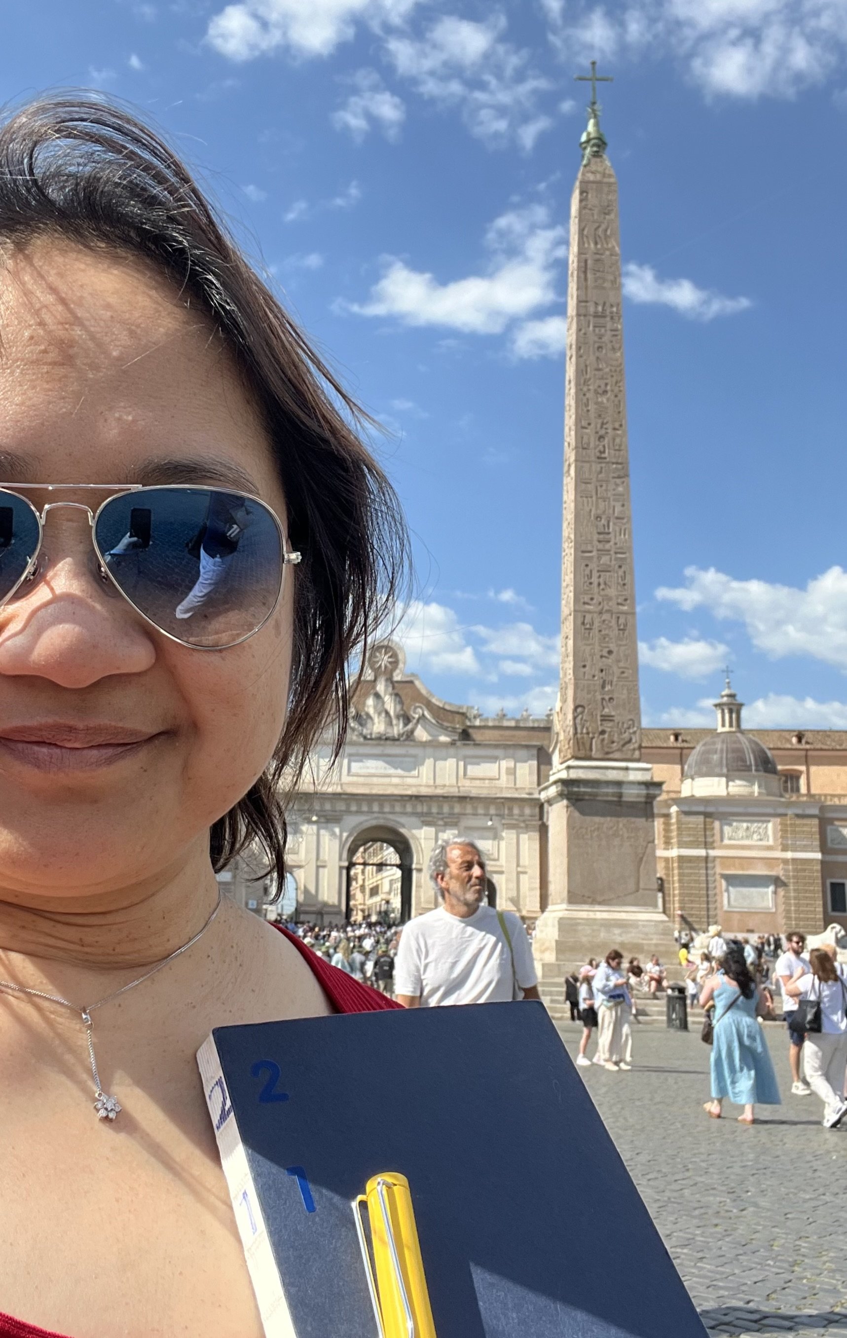 A woman wearing sunglasses and a red top taking a selfie at St. Peter's Square in Vatican City, with the Egyptian obelisk and basilica in the background, on a sunny day with a blue sky and clouds.