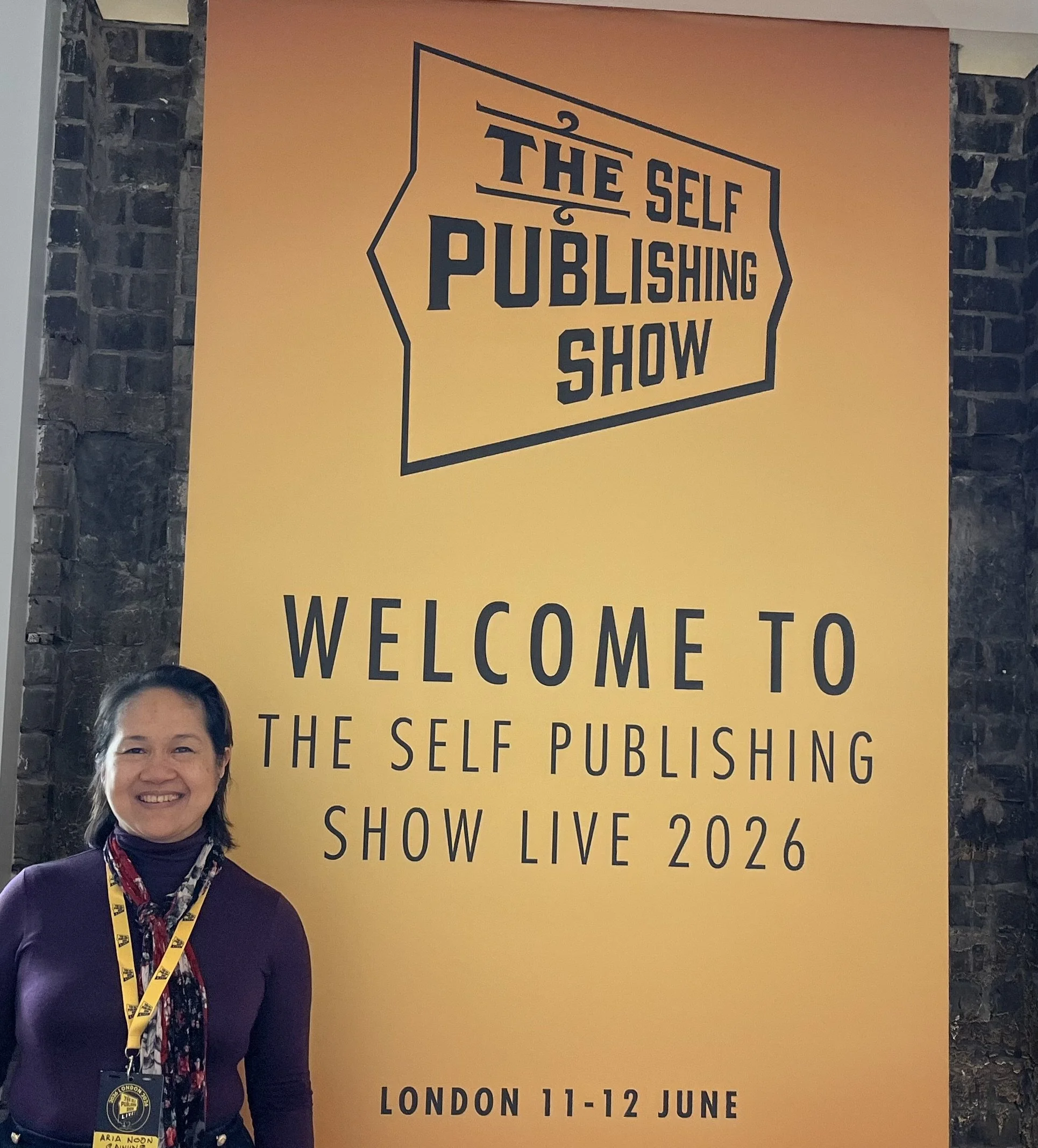 A woman smiling, wearing a dark purple top, a patterned scarf, and a London Pubsign live 2022 badge on a lanyard. Behind her is a large orange banner with black text about a self-publishing show happening in London on June 11-12.