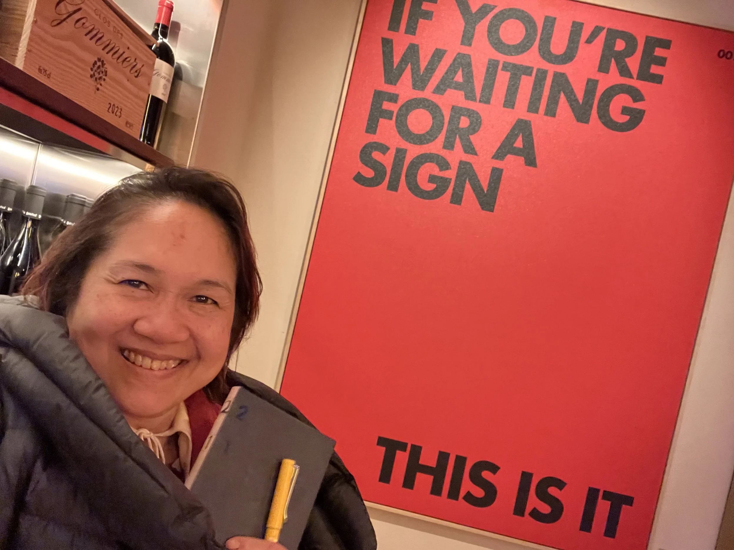 A woman smiling in a black jacket with a brown shirt underneath, holding a yellow pen and a smartphone, standing in front of a large red sign with black and gray text that says, 'IF YOU'RE WAITING FOR A SIGN THIS IS IT.' In the background, there are 