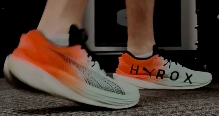 Close-up of a person wearing orange and white running shoes, with the word 'HYROX' printed on the side of one shoe.