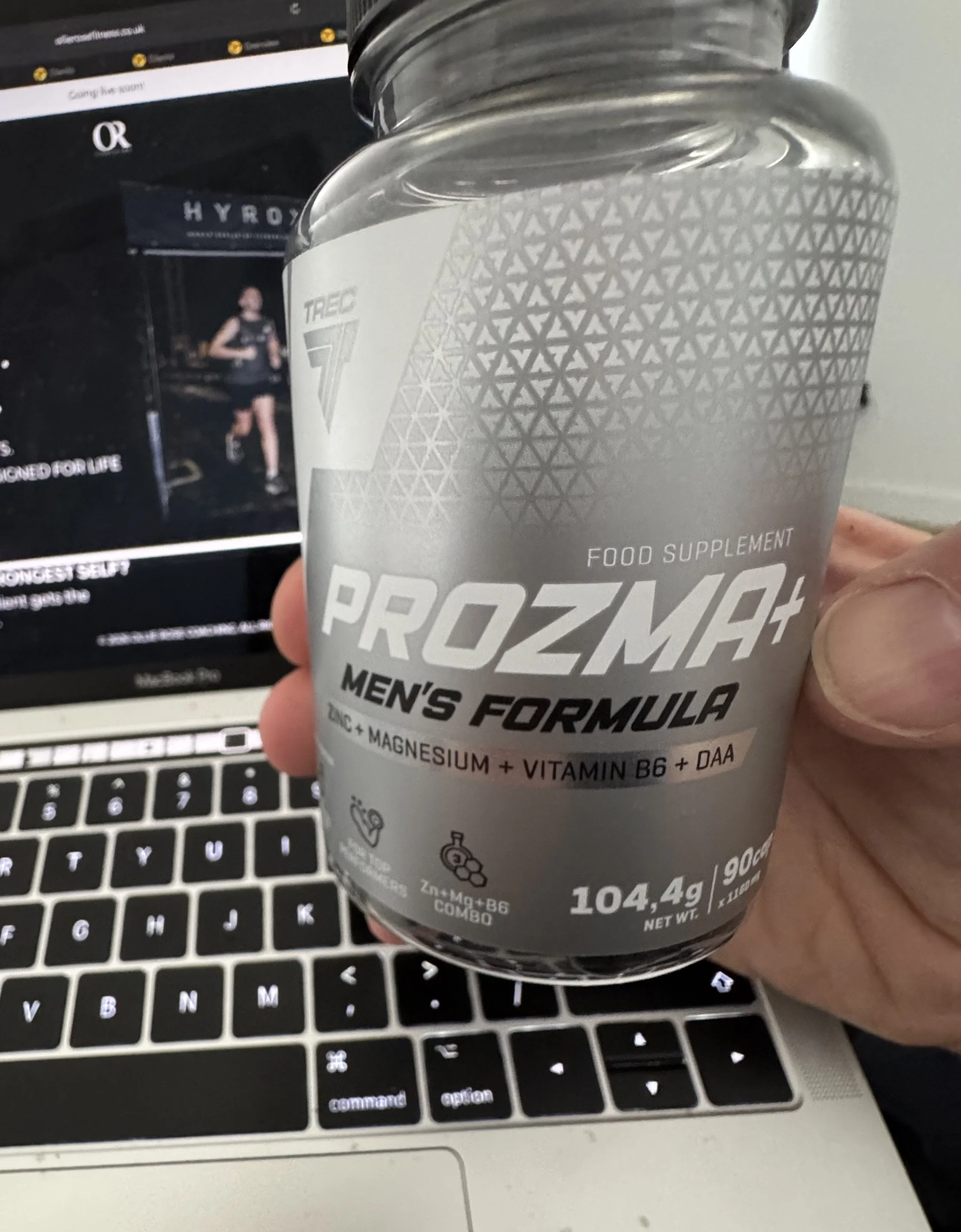 A person holding a gray container of Prozymes men's formula food supplement with a background of a laptop keyboard and a blurred screen showing a woman running.