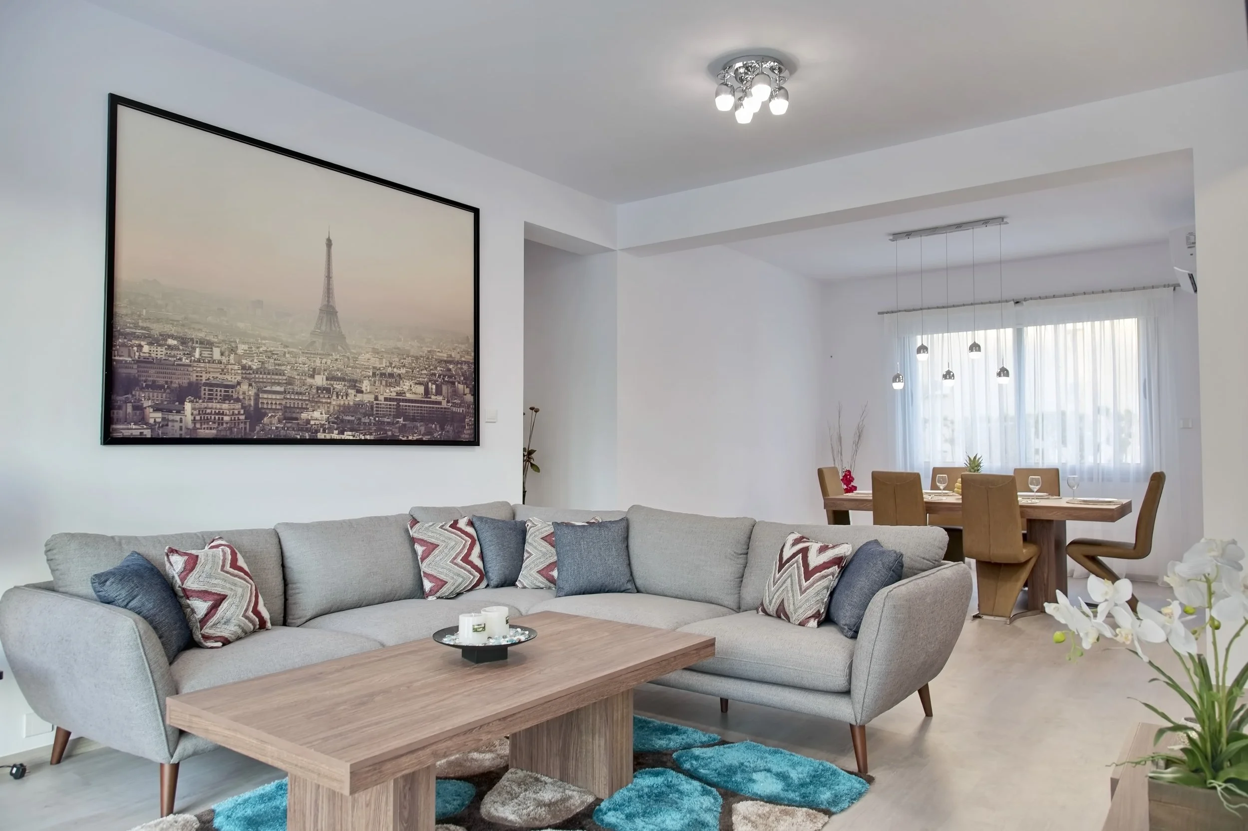 A living room with a light gray sofa, colorful patterned pillows, a wooden coffee table, a large wall art of Paris with the Eiffel Tower, a dining table with chairs, and a bright window with white curtains.