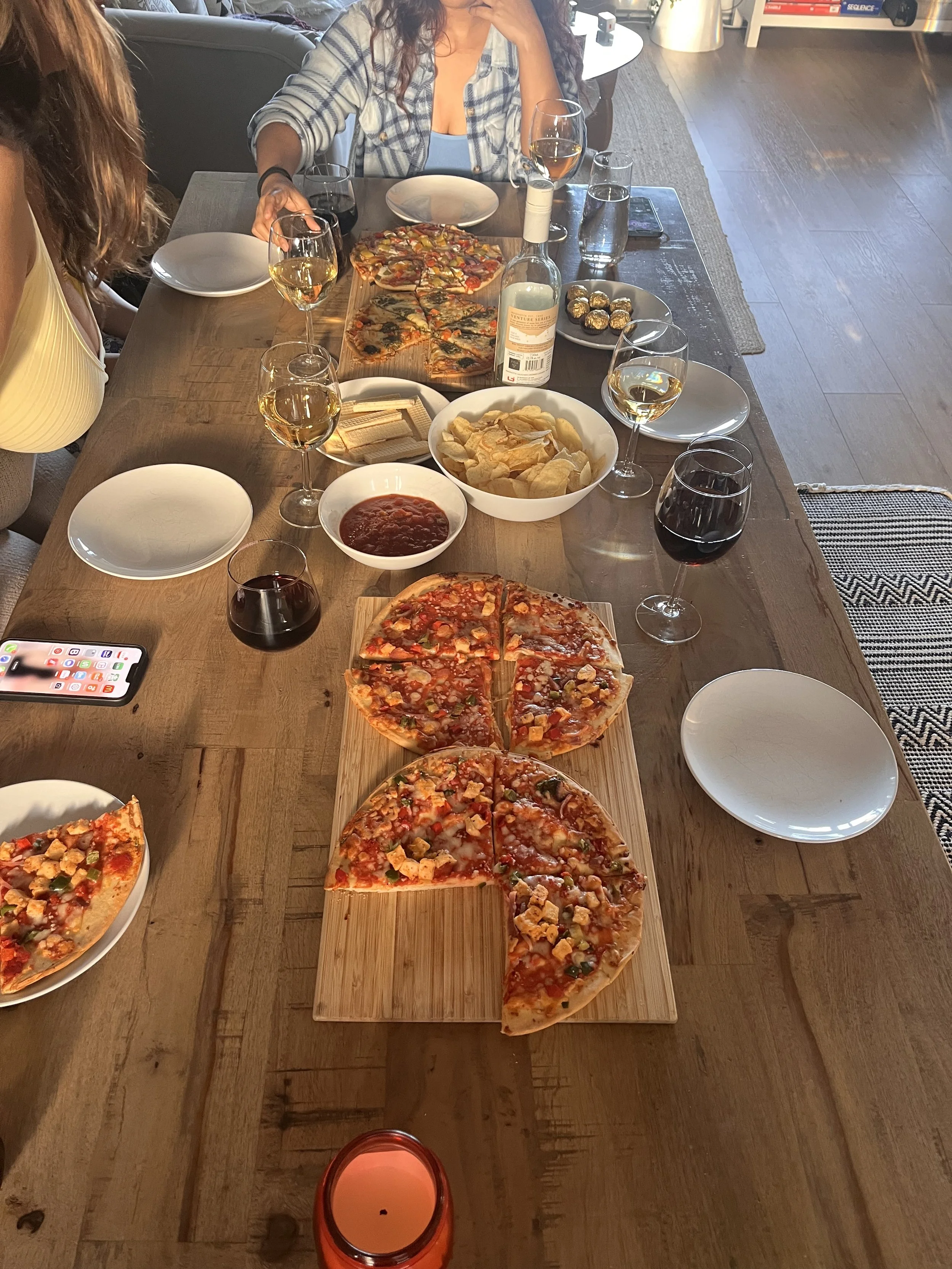 A dining table with various food and drinks including pizza, chips, wine, and chocolate. People sitting around with wine glasses and plates. A smartphone is visible on the table.