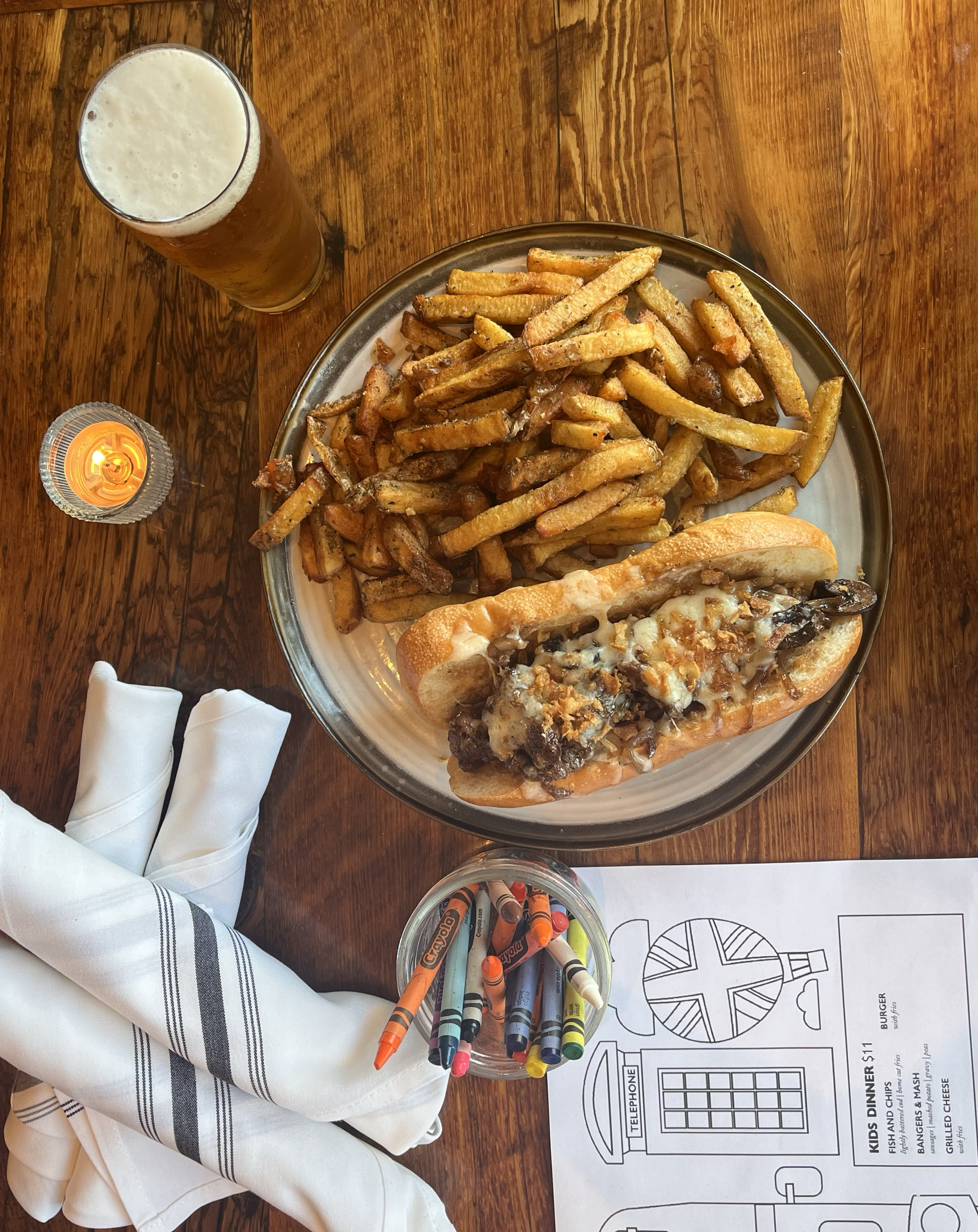 A plate of French fries and a cheesesteak sandwich on a wooden table, accompanied by a pint of beer, a lit candle, and rolled napkins. There is a children's coloring sheet and a jar of crayons next to the plate.