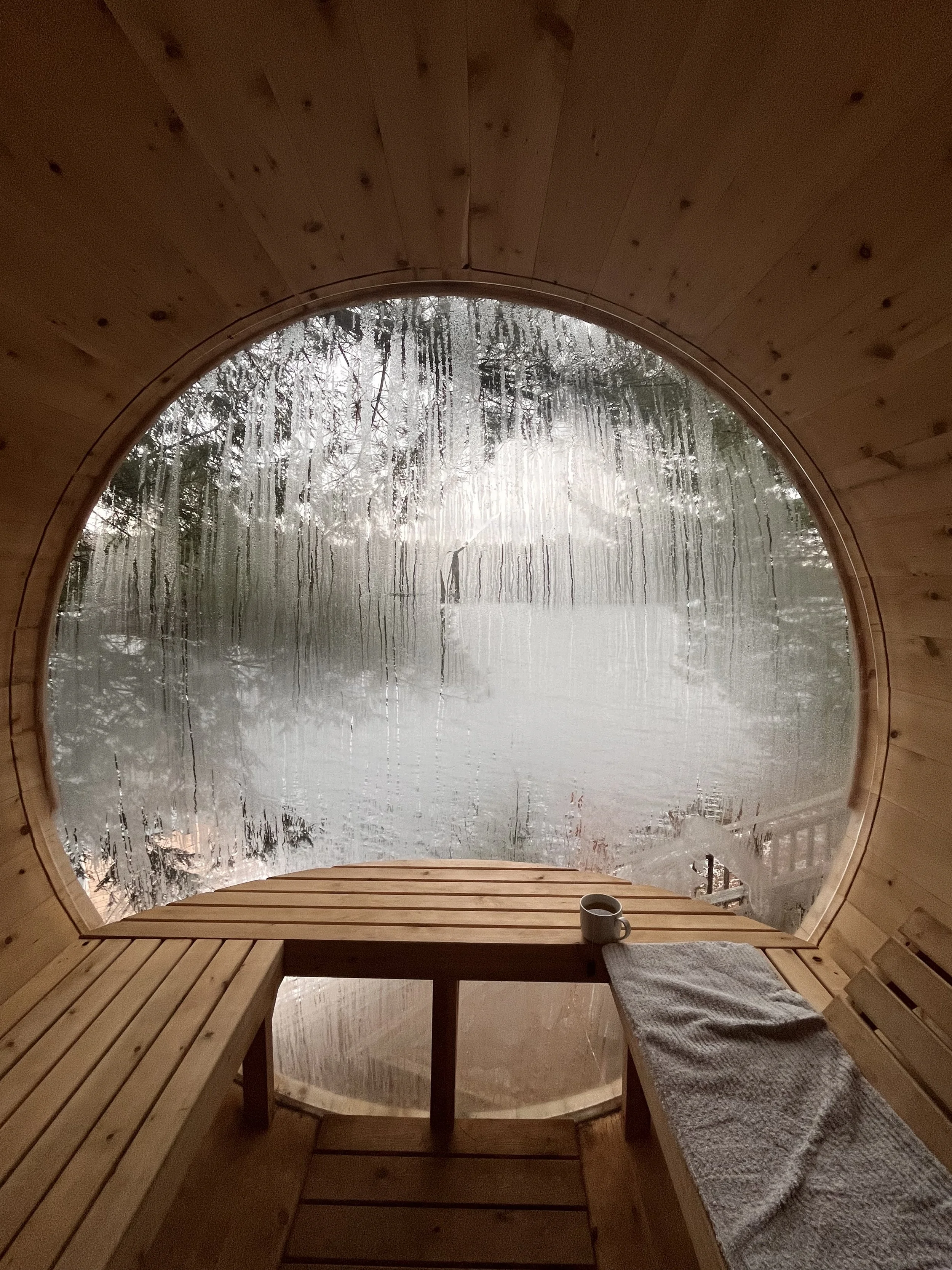 Interior of a wooden barrel sauna with a foggy glass window, a bench, and a towel alongside a cup on the seat.