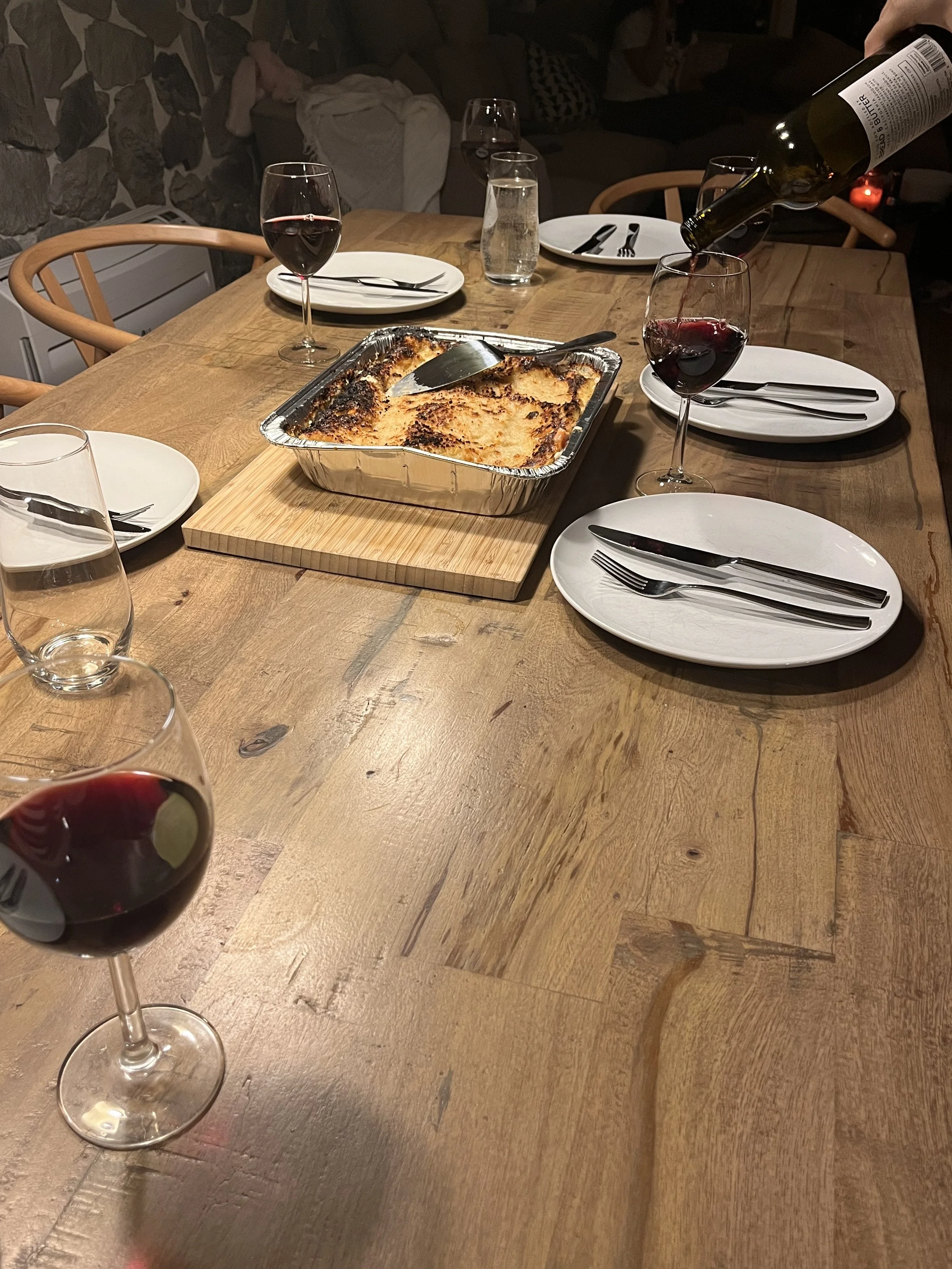Table set with lasagna in foil tray, plates, cutlery, wine glasses, and being served with red wine.