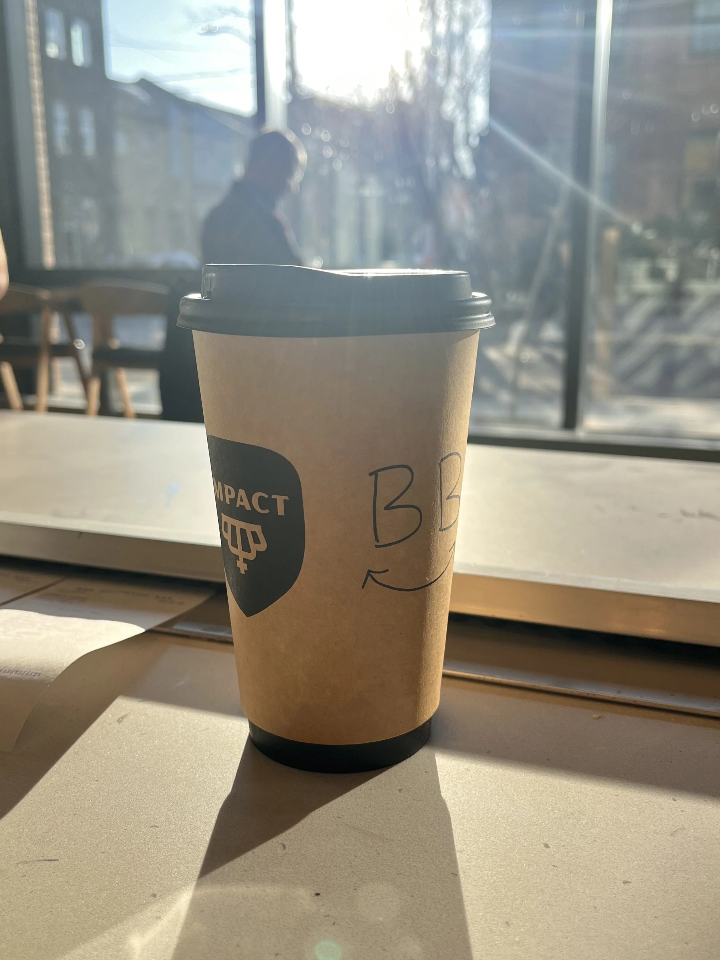 A paper coffee cup with a lid and a handwritten "BB" on it, placed on a table with sunlight streaming through a window in a cafe setting.