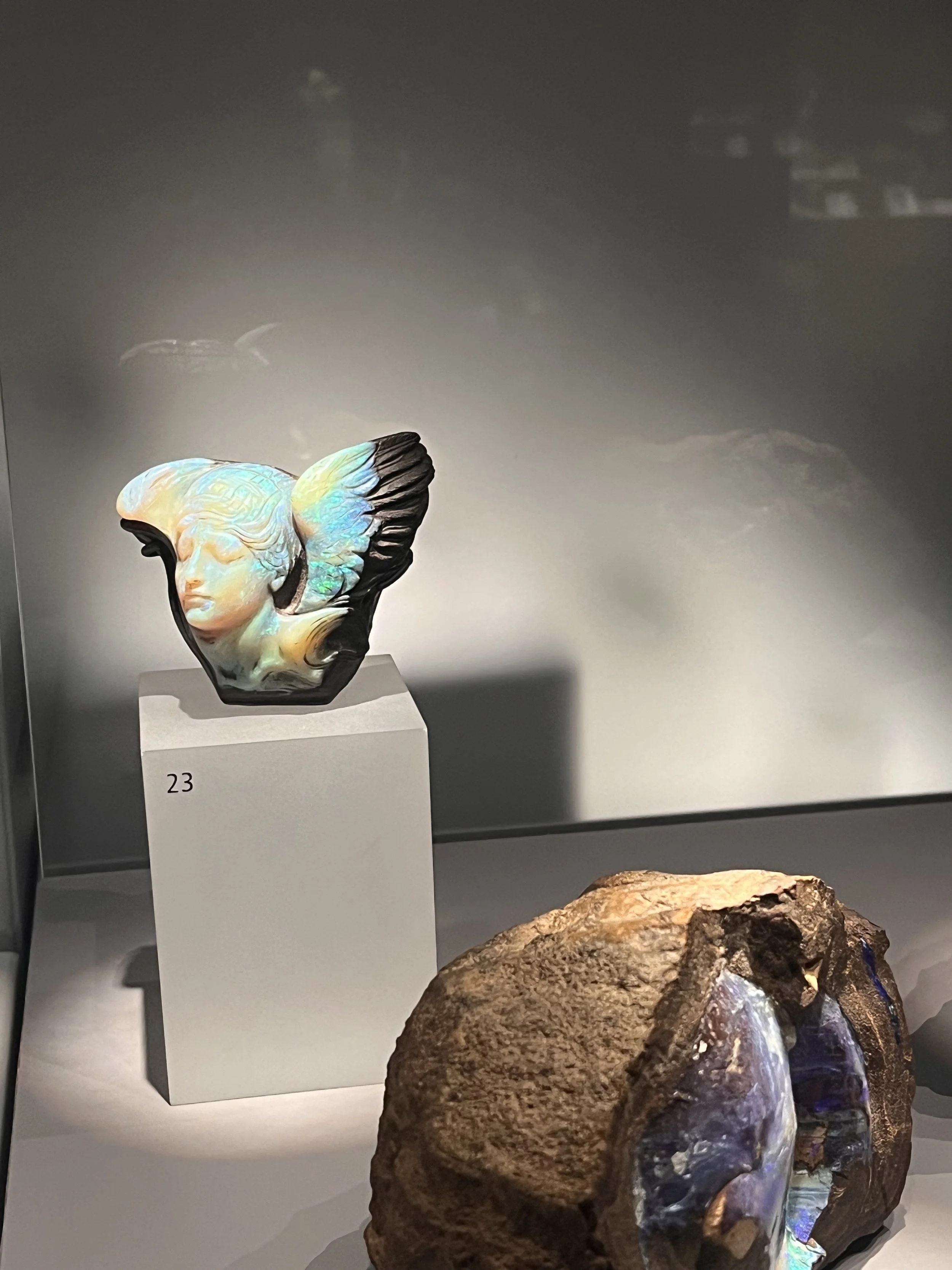 An opal sculpture depicting a human face with wings, displayed on a pedestal numbered 23, alongside a large opal rock in a museum exhibition ambiance.