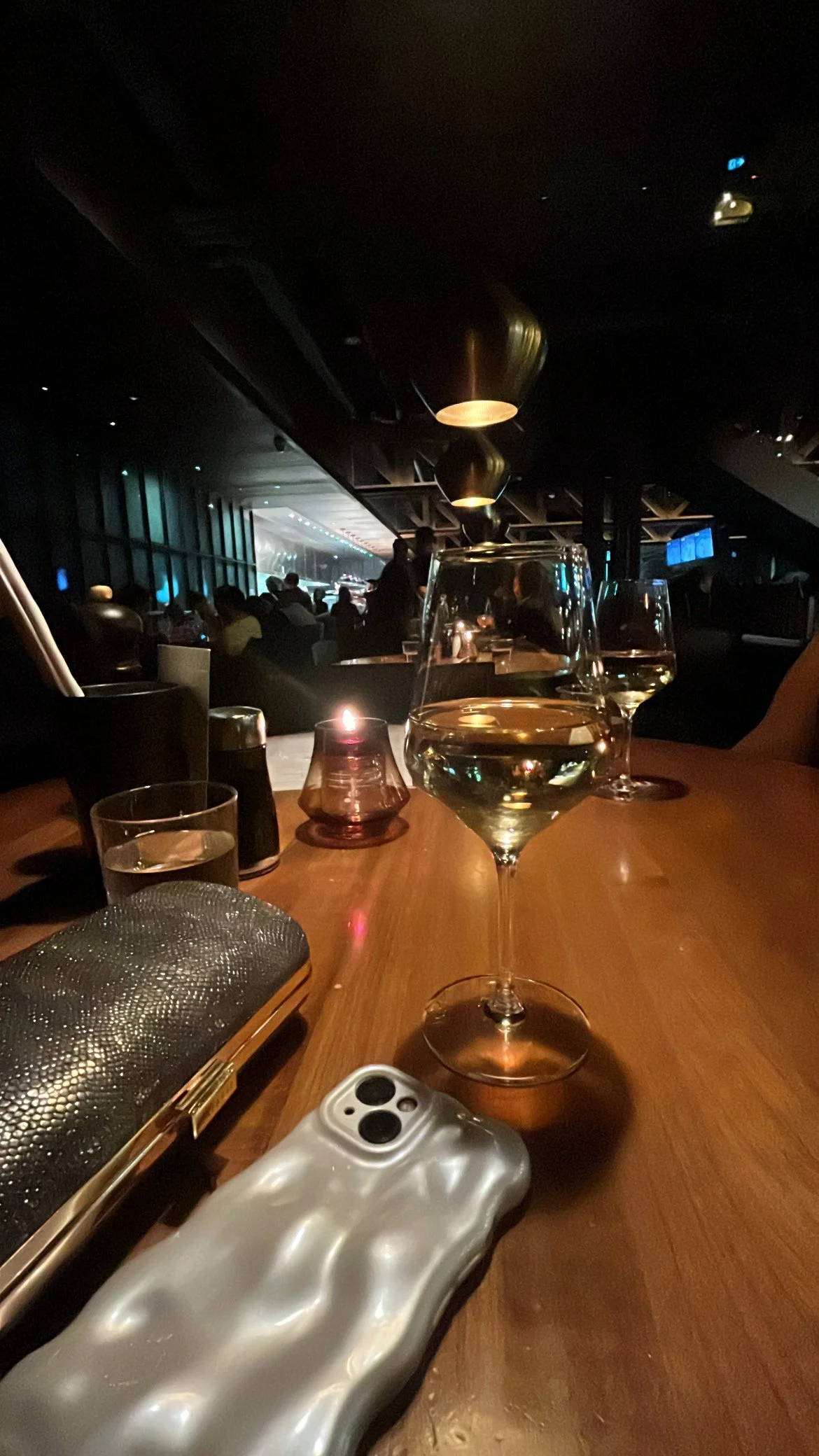 Dimly lit modern restaurant interior with wine glasses, a lit candle, a clutch, and a phone on a wooden table.