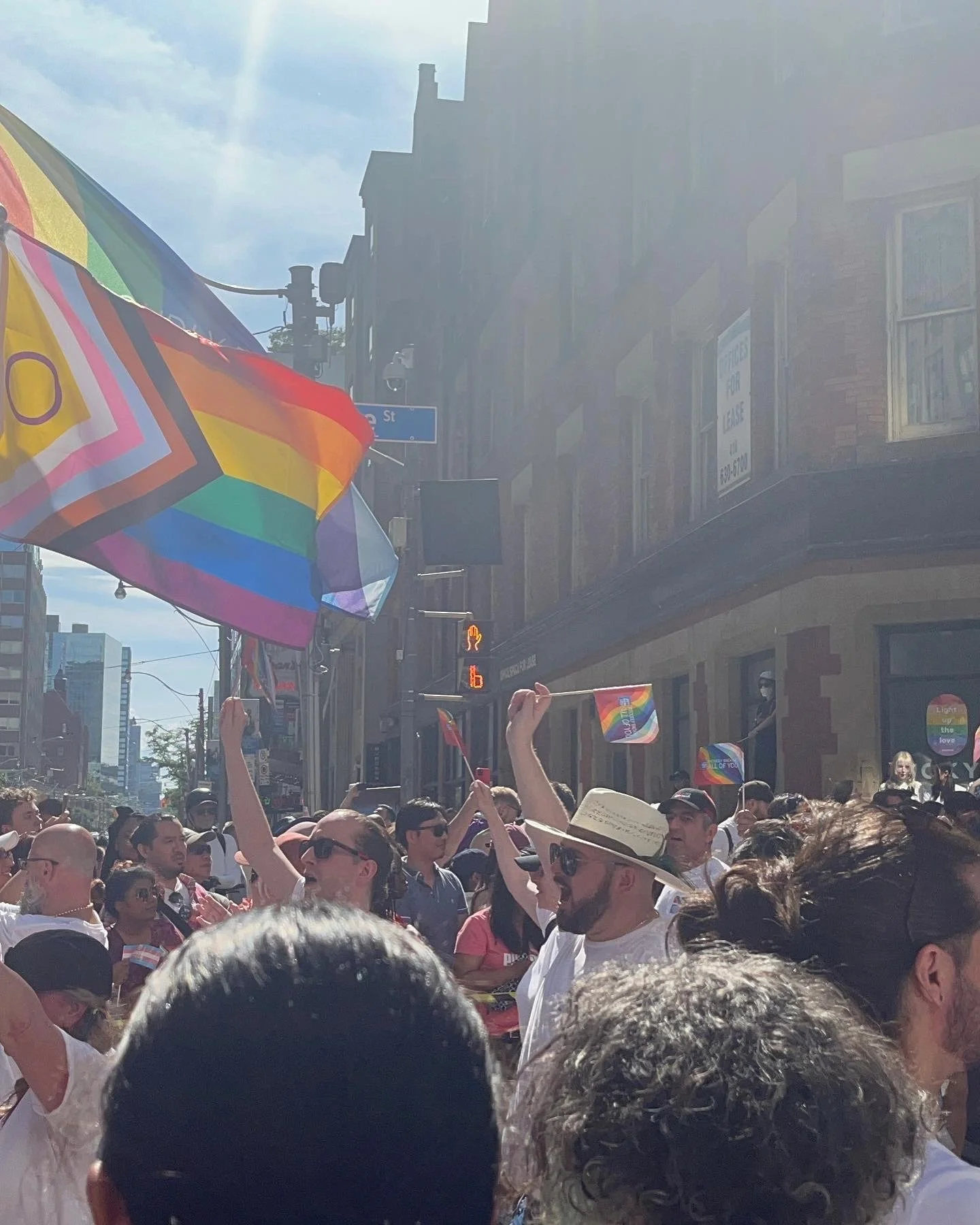People at a pride parade holding colorful LGBTQ+ flags on a city street with buildings and traffic signal visible; sunny day.