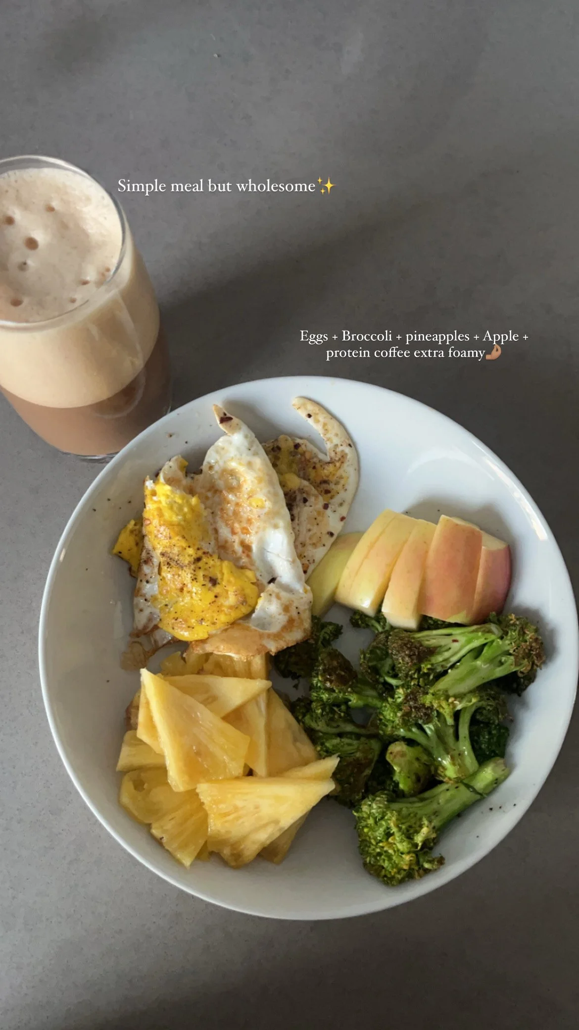 Plate with eggs, broccoli, pineapple, apple slices, and a glass of foamy protein coffee.