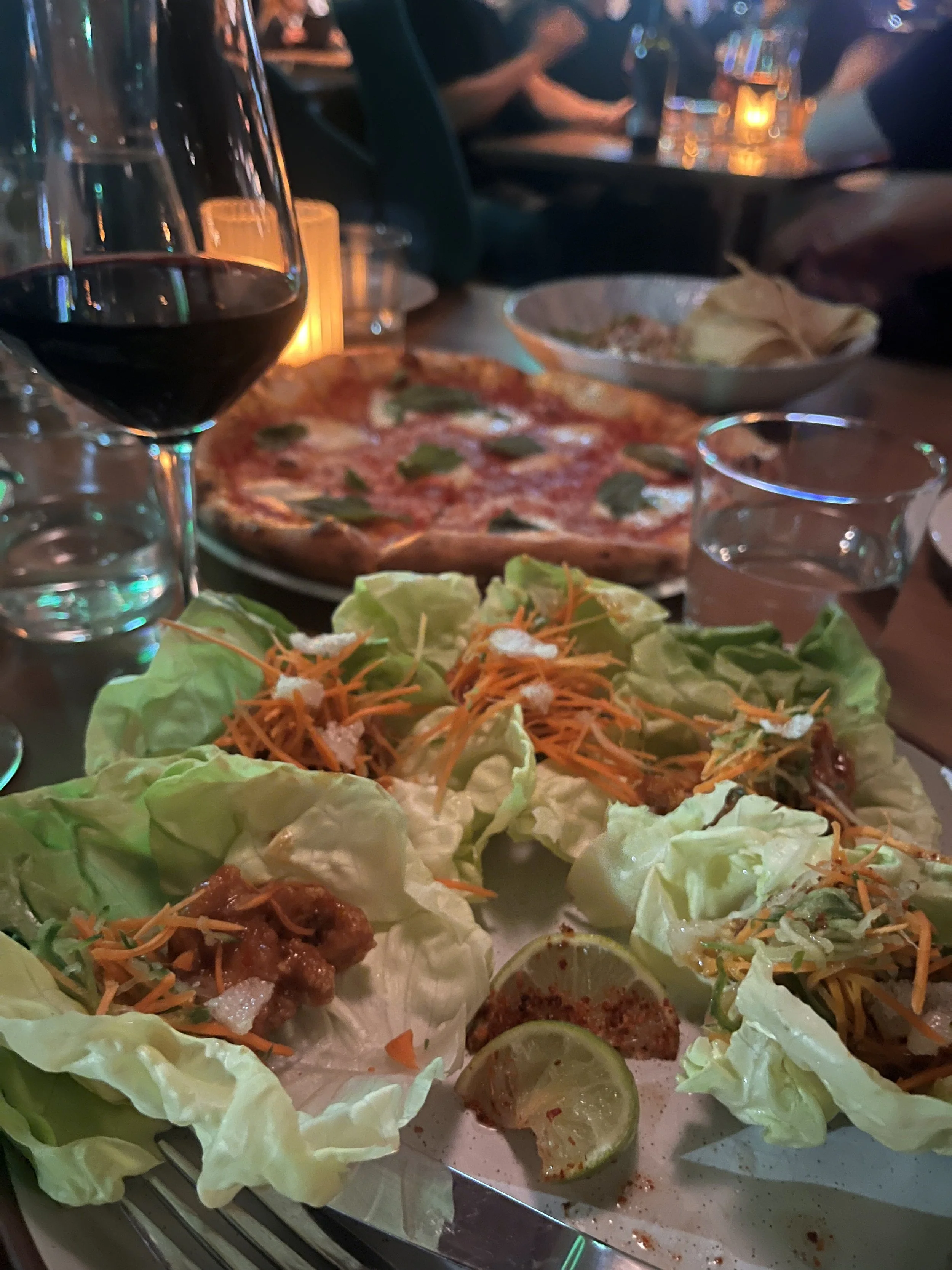 A table with lettuce wraps featuring shredded carrots and a small amount of sauce, lime wedges, and a glass of red wine in the foreground. A pizza topped with basil is in the background, along with a bowl of chips.