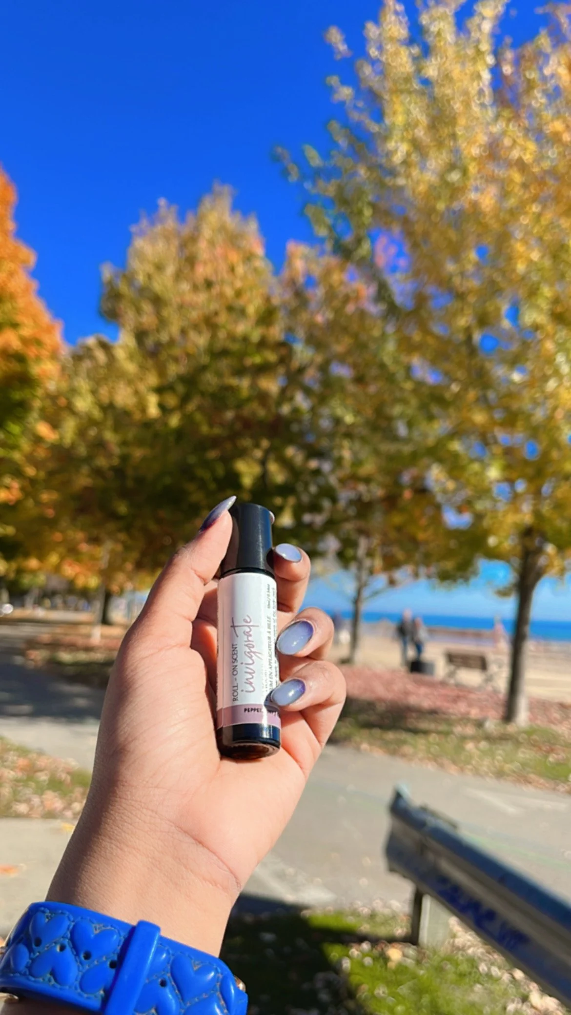 Hand holding a roll-on scent bottle labeled 'Invigorate' against a background of autumn trees and a clear blue sky.