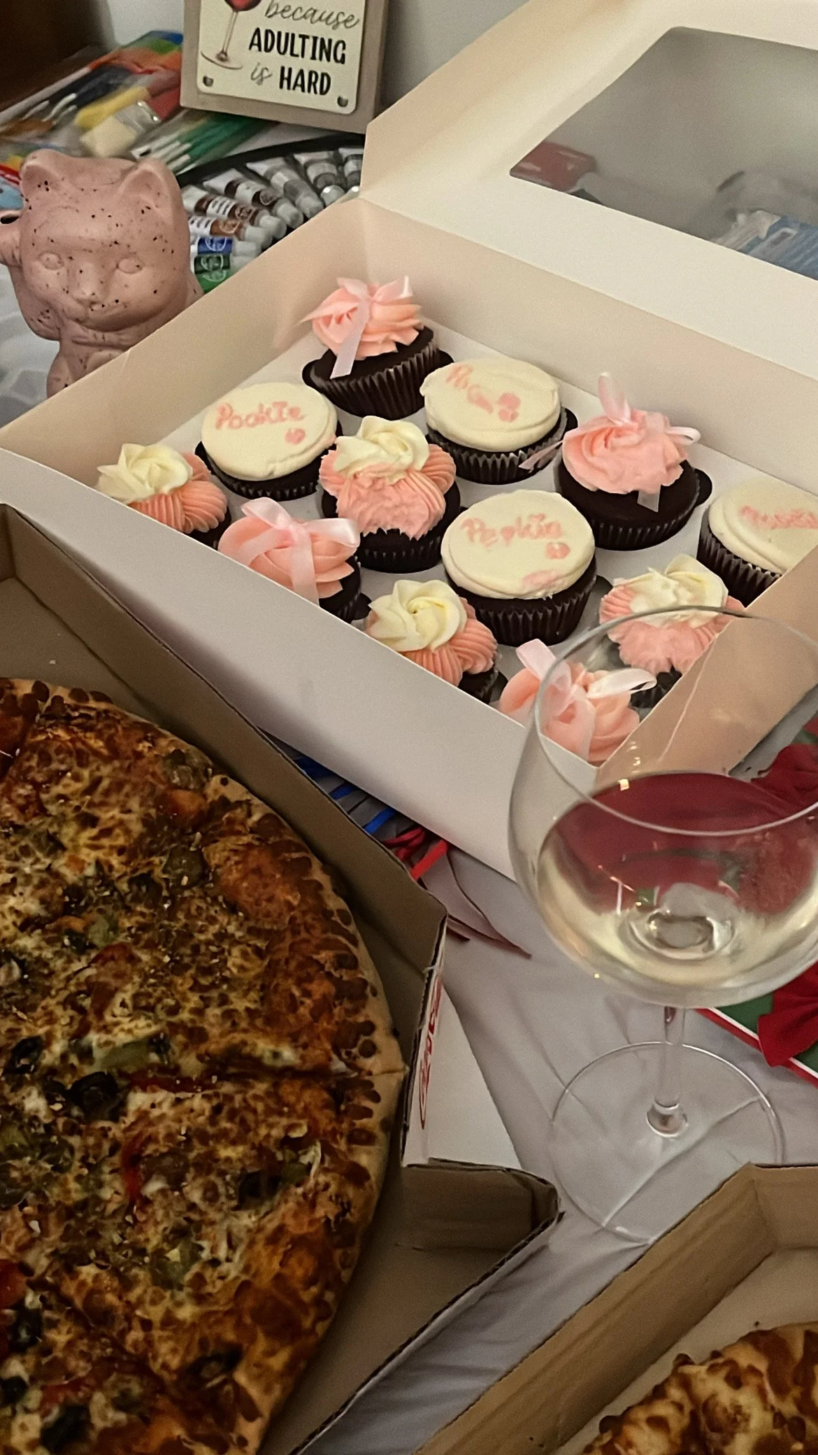 Box of cupcakes with pink and white frosting, a pizza in a box, a wine glass, and art supplies in the background.