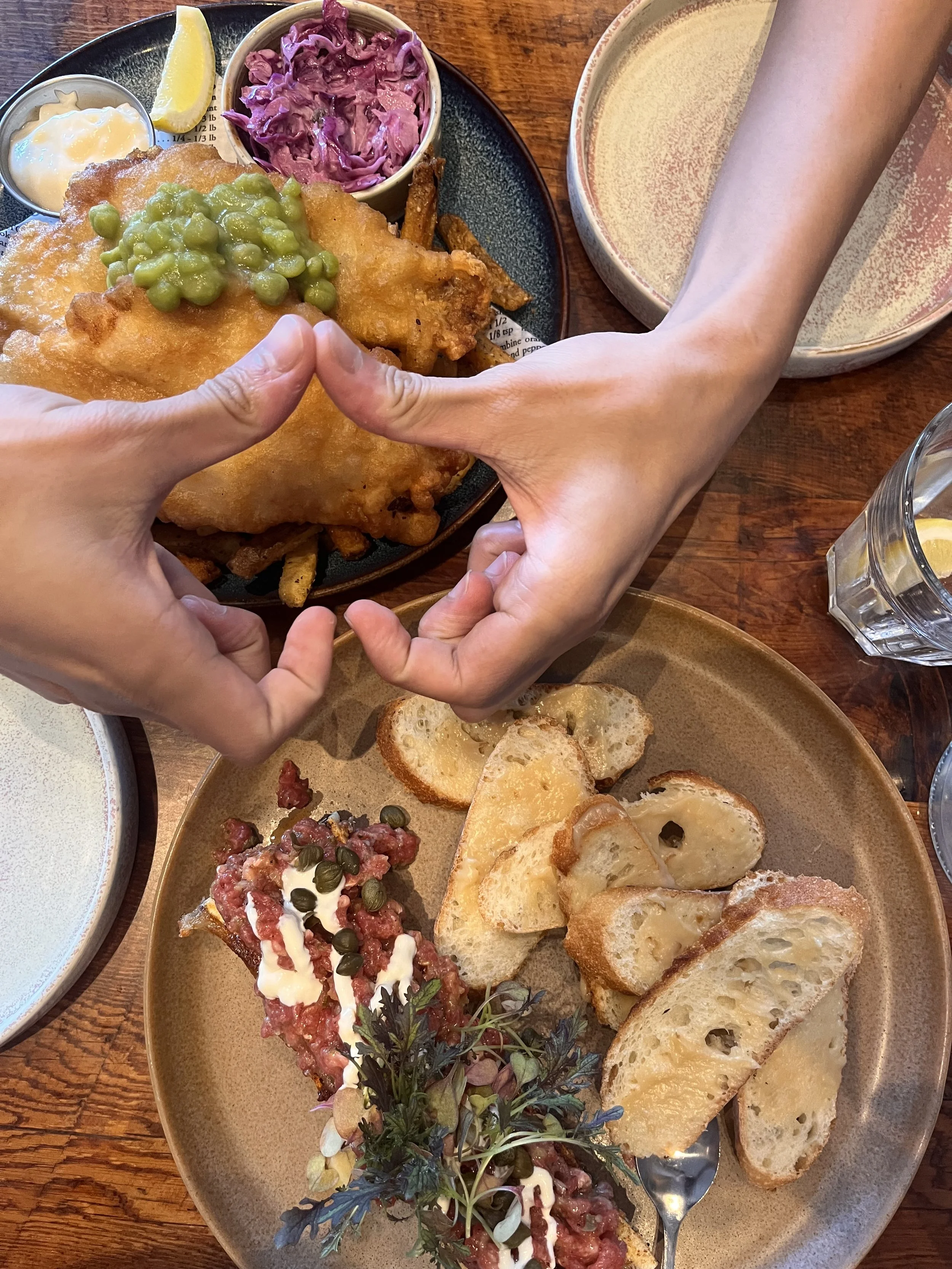 Fish and chips with mushy peas, purple coleslaw, and tartar sauce on one plate; beef tartare with capers, cream sauce, and herbs served with slices of bread on another plate; hands forming a shape in the foreground.