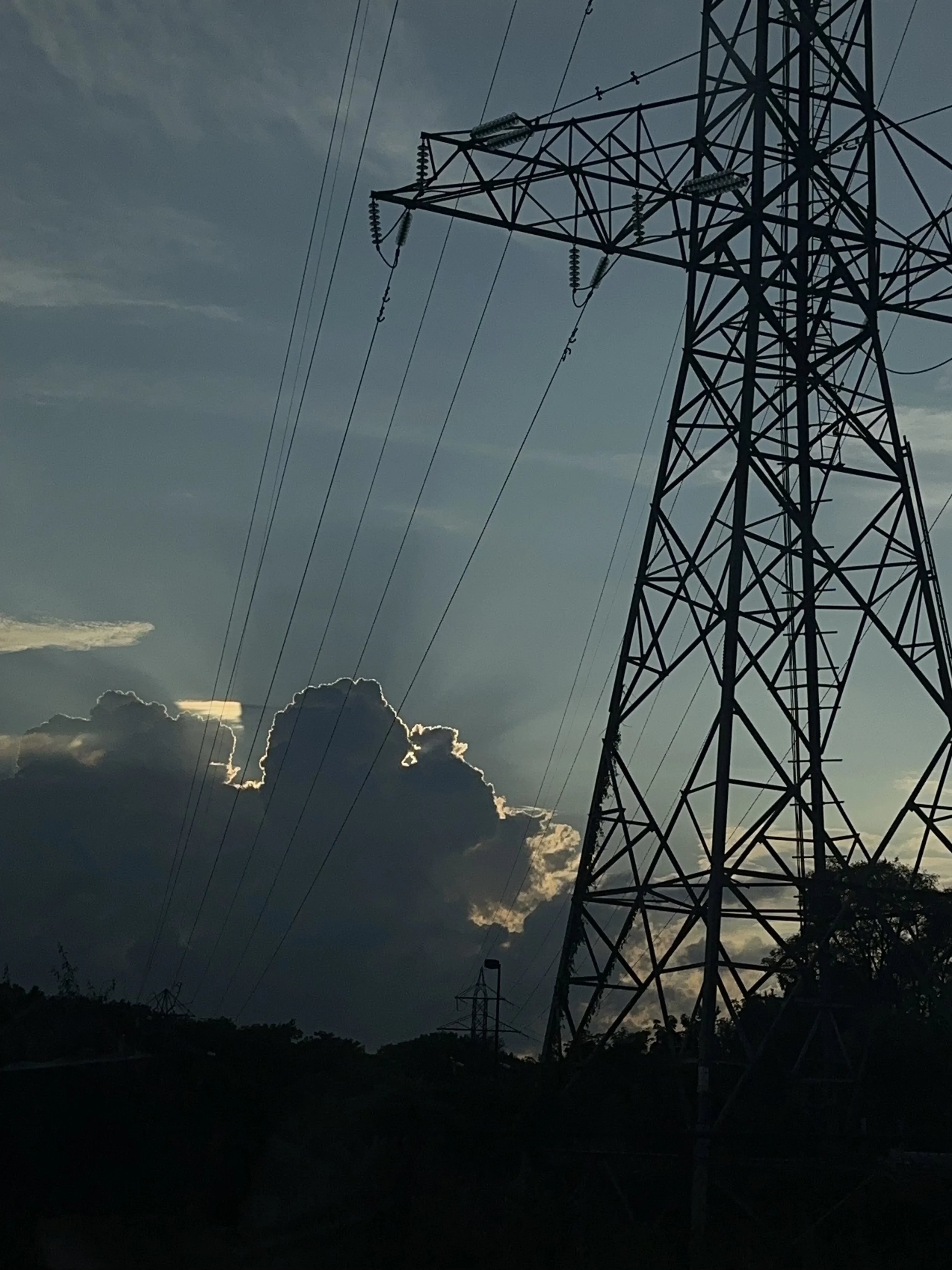 Silhouette of a power line tower with cables against a cloudy sky at sunset.
