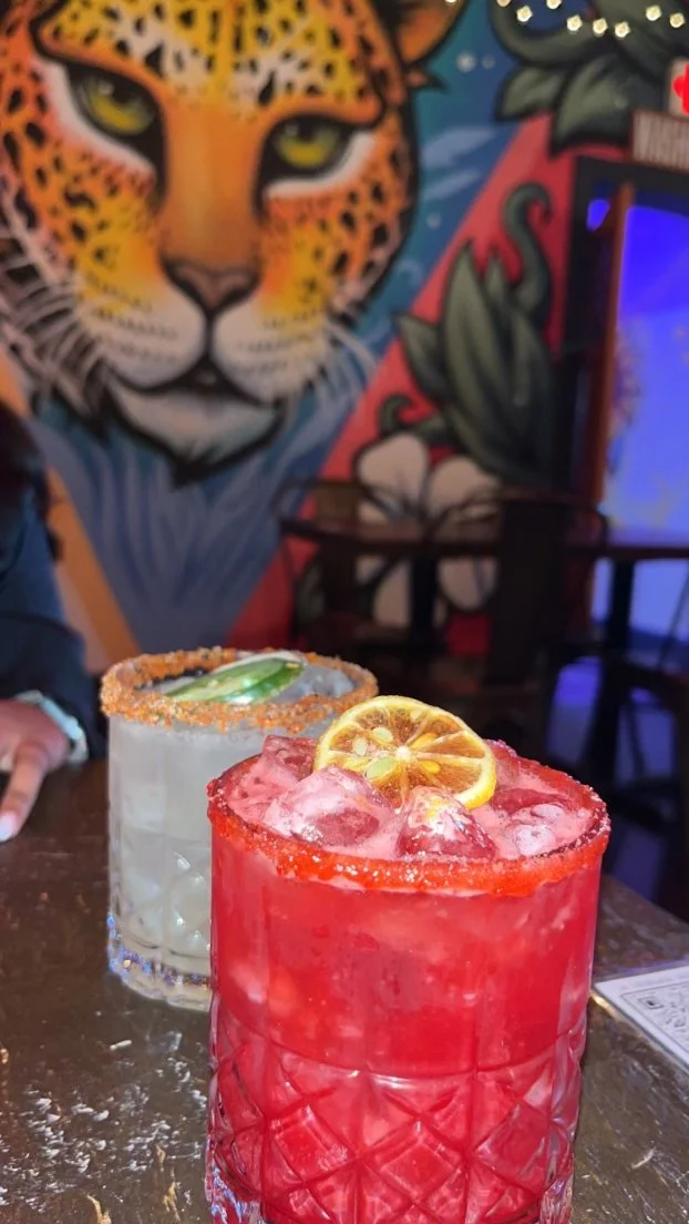 Two colorful cocktails on a table with a vibrant mural of a leopard in the background.