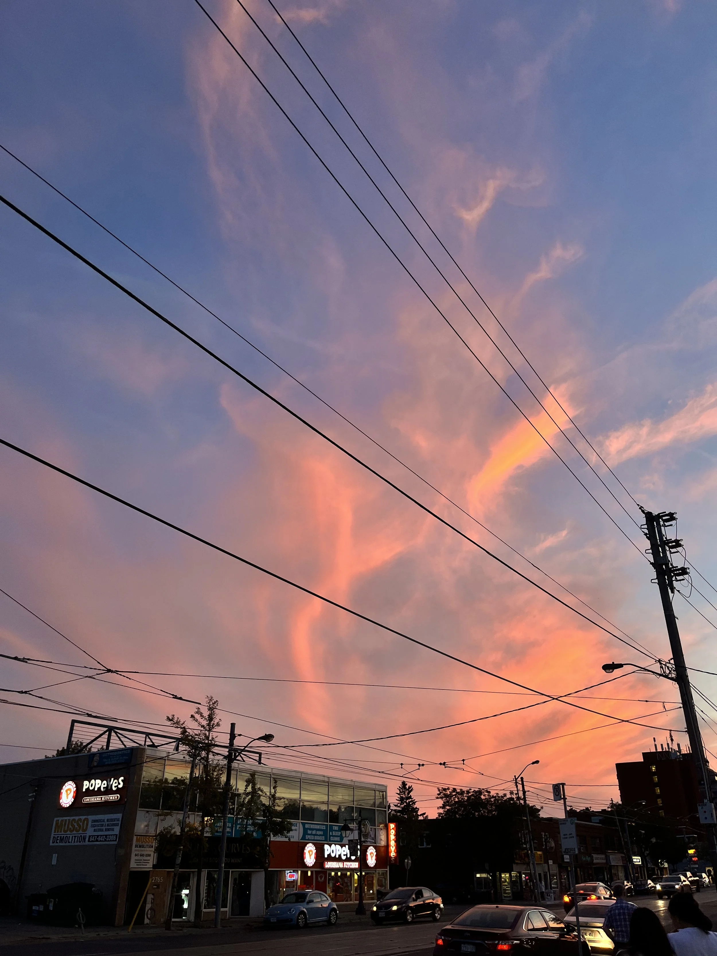 Evening sky with pink clouds over a city street, power lines overhead, and a Popeyes restaurant in the foreground.