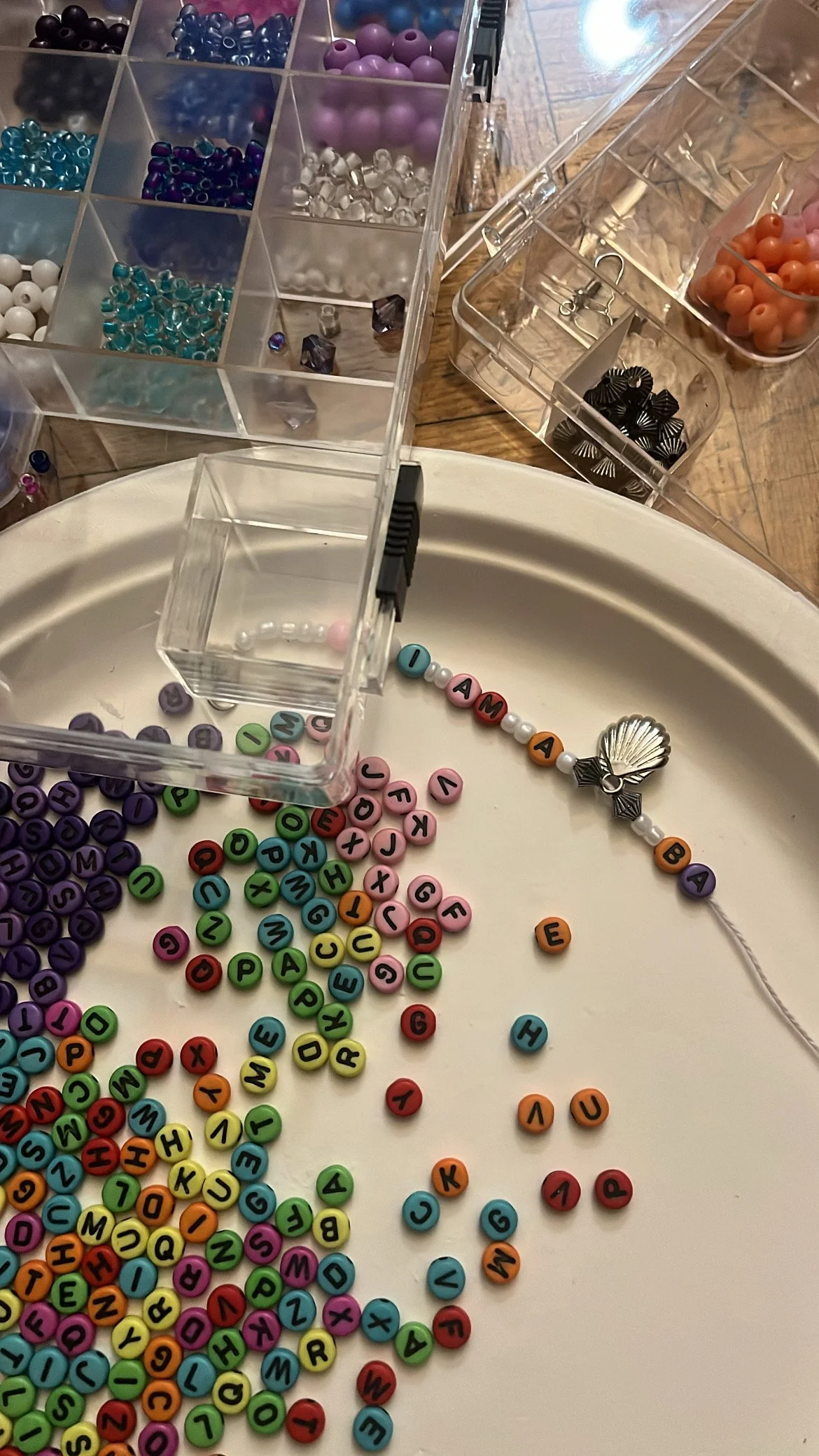Crafting supplies including colorful alphabet beads, assorted beads in a divided container, and a string with a shell charm on a plate.
