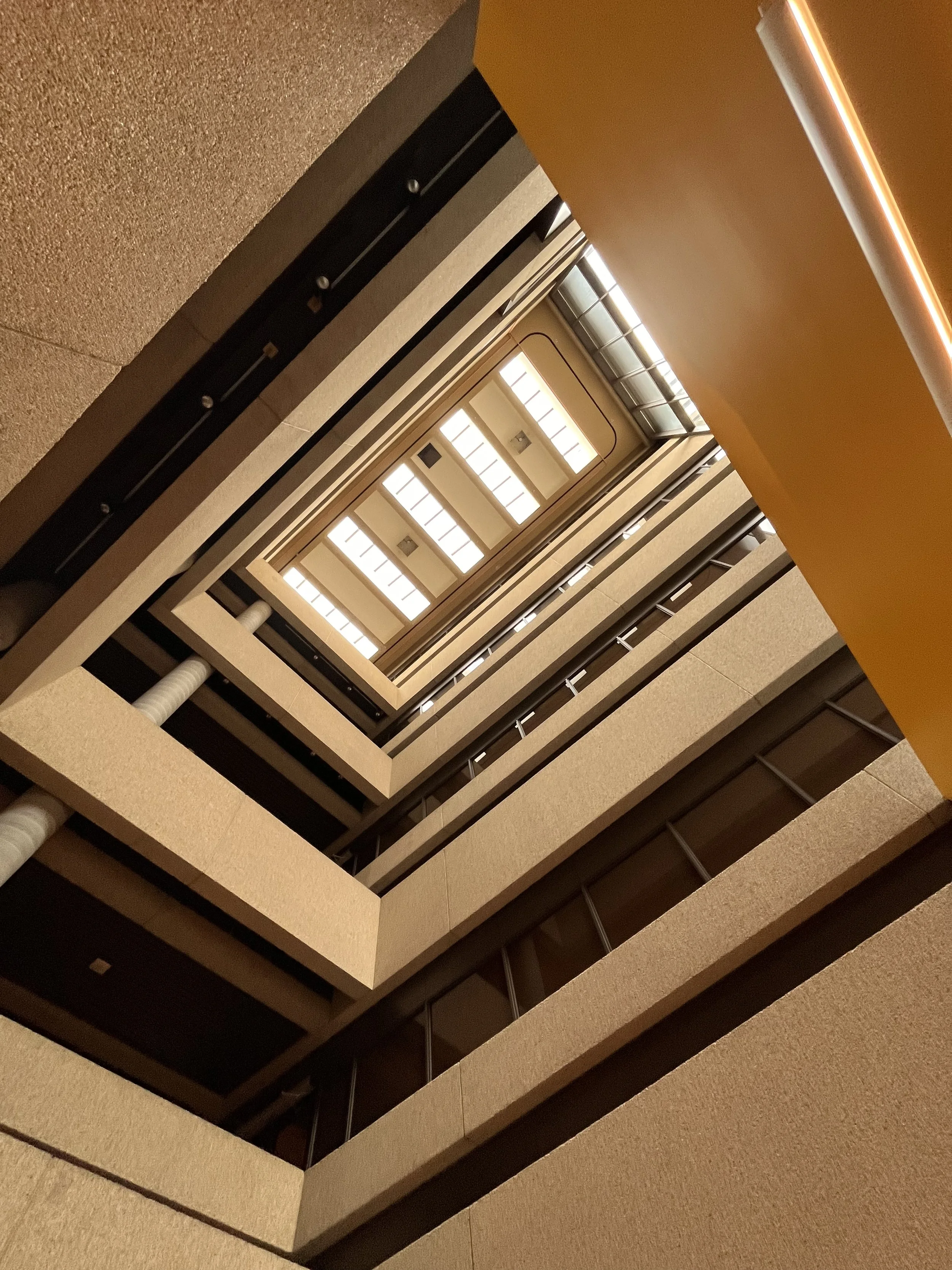 Looking up the inside of a multi-story building atrium with symmetrical balconies and a bright ceiling light."}