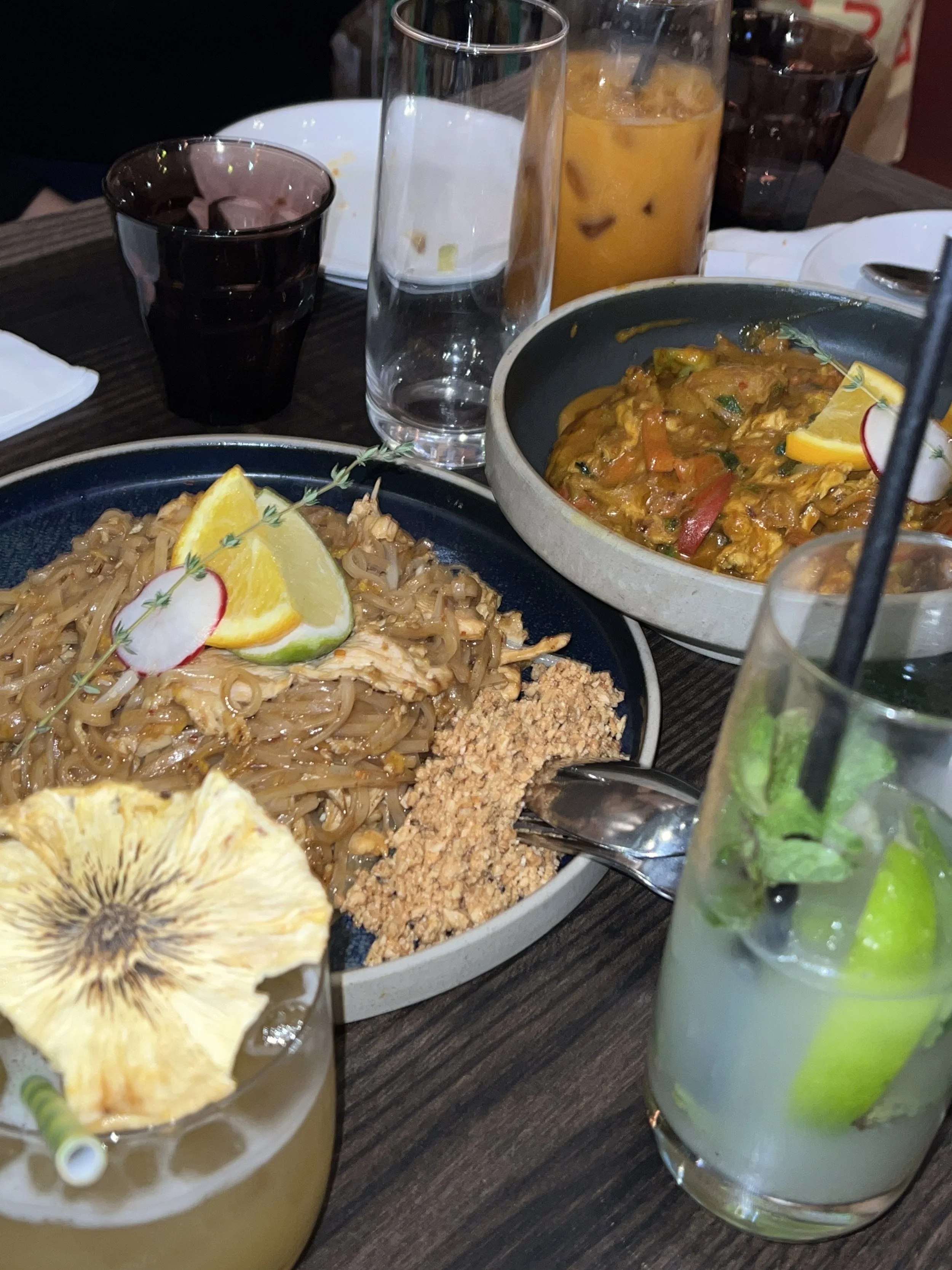Table with plates of pad Thai and curry, surrounded by drinks including a cocktail with a dried pineapple garnish and a glass of iced tea.