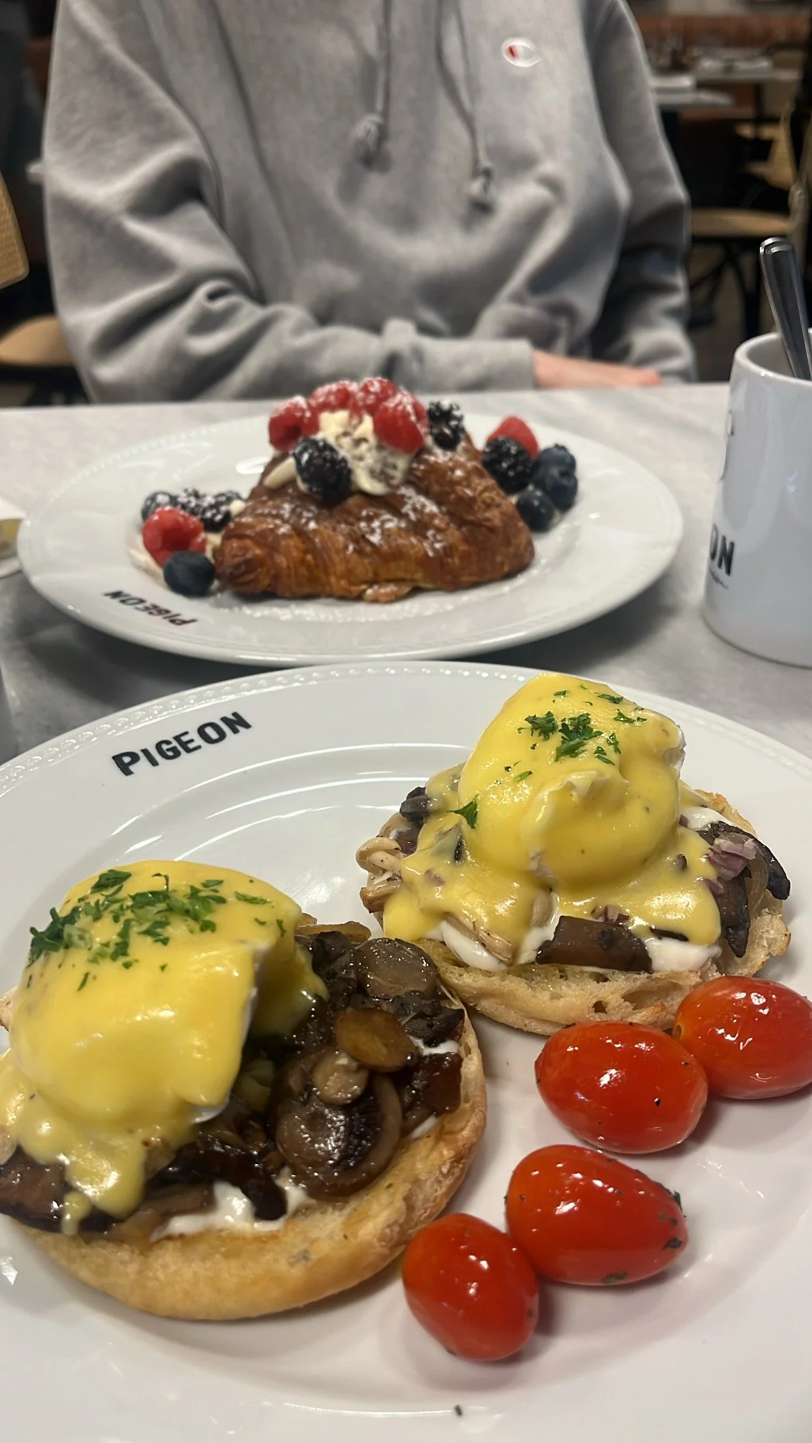 Close-up of a breakfast dish featuring eggs Benedict with mushrooms and cherry tomatoes, and a croissant topped with berries and cream, served on 'Pigeon' branded plates. A person in a gray hoodie is blurred in the background.