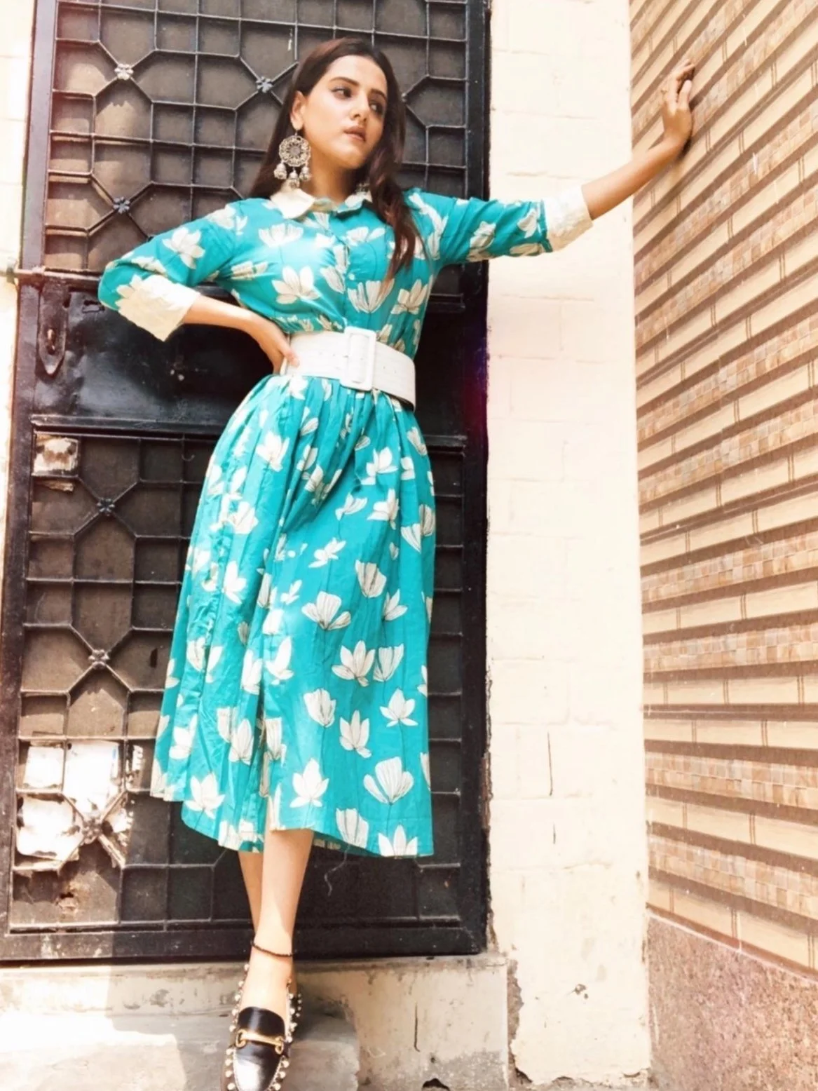 Woman posing against a patterned wall, wearing a teal dress with white flowers and a white belt, accessorized with large earrings and black loafers.