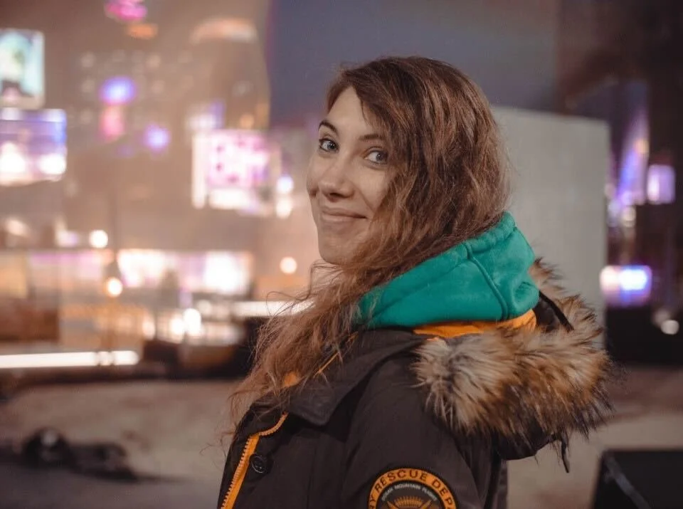 A young woman with long, wavy brown hair wearing a black jacket with a fur-lined hood, a teal hoodie underneath, and a Rescue Department patch on her sleeve, standing outdoors at night with city lights in the background.