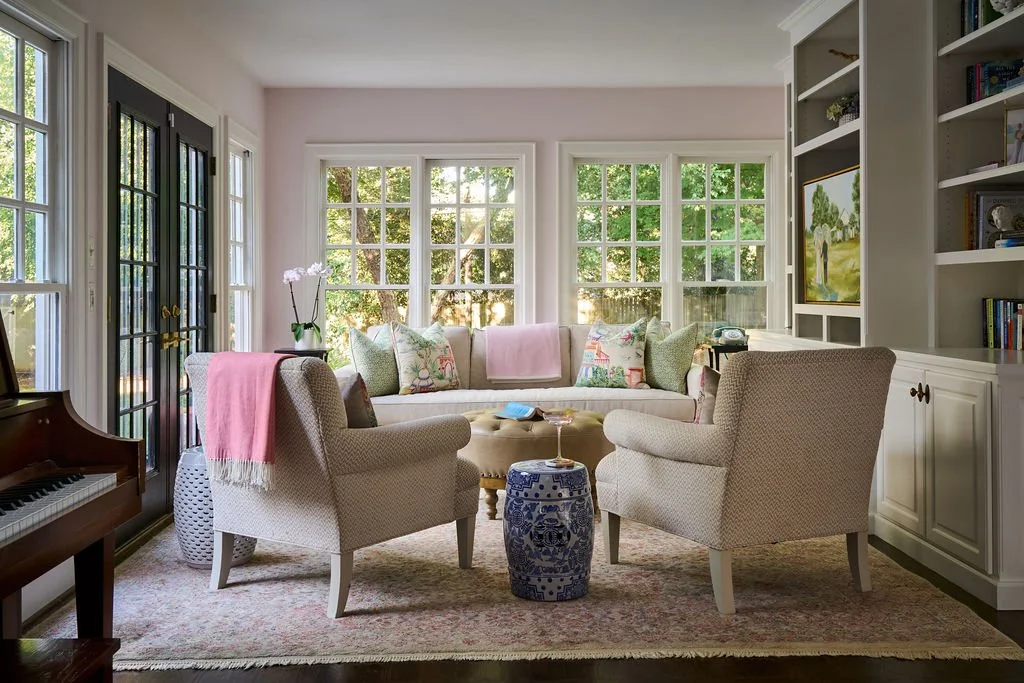 Bright sunroom with large windows, a sofa with colorful pillows, two armchairs, a chinoiserie garden stool turned side table with white built-ins filled with books, family heirlooms. The room is painted the lightest hue of pink offering a warm glow.