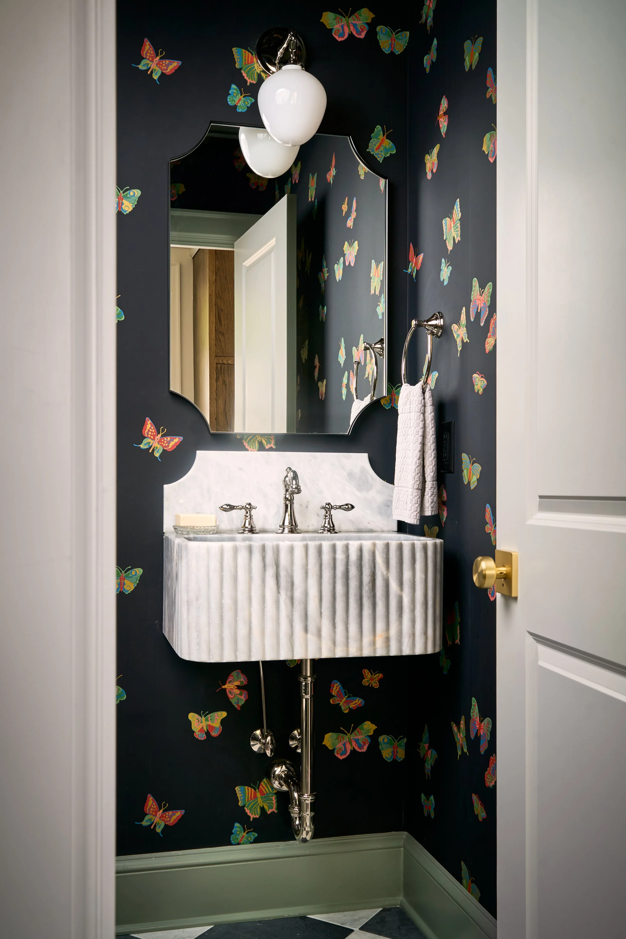 A bathroom mirror with a black background decorated with colorful butterfly wallpaper. There are white sconce lights, a white hand towel hanging on a chrome ring, and a marble sink with chrome fixtures.