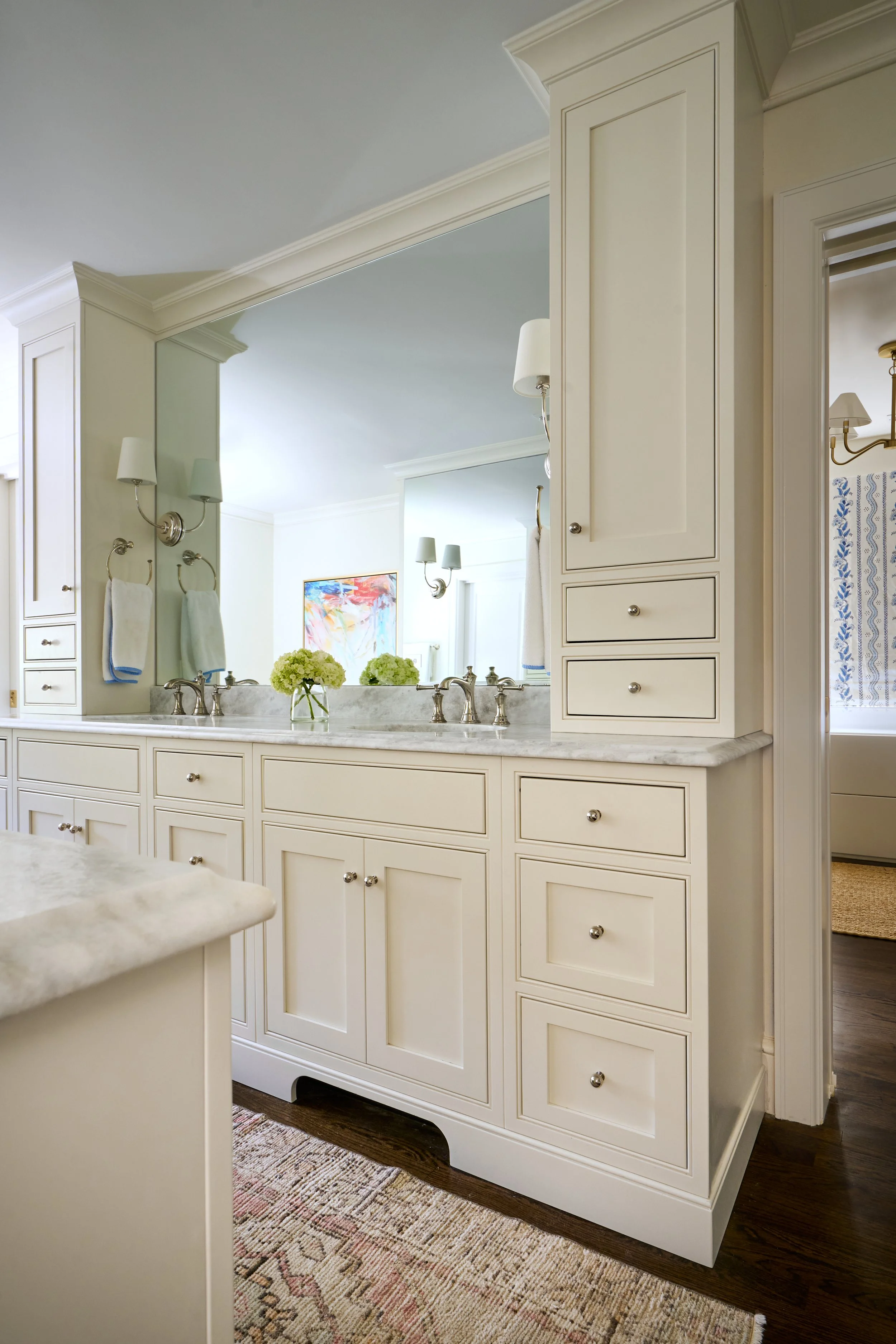 Primary bathroom with off white colored custom inset cabinetry featuring two vanity towers, an oversized vanity mirror, two visual comfort sconces, beautiful marble countertops and dark wood floors.