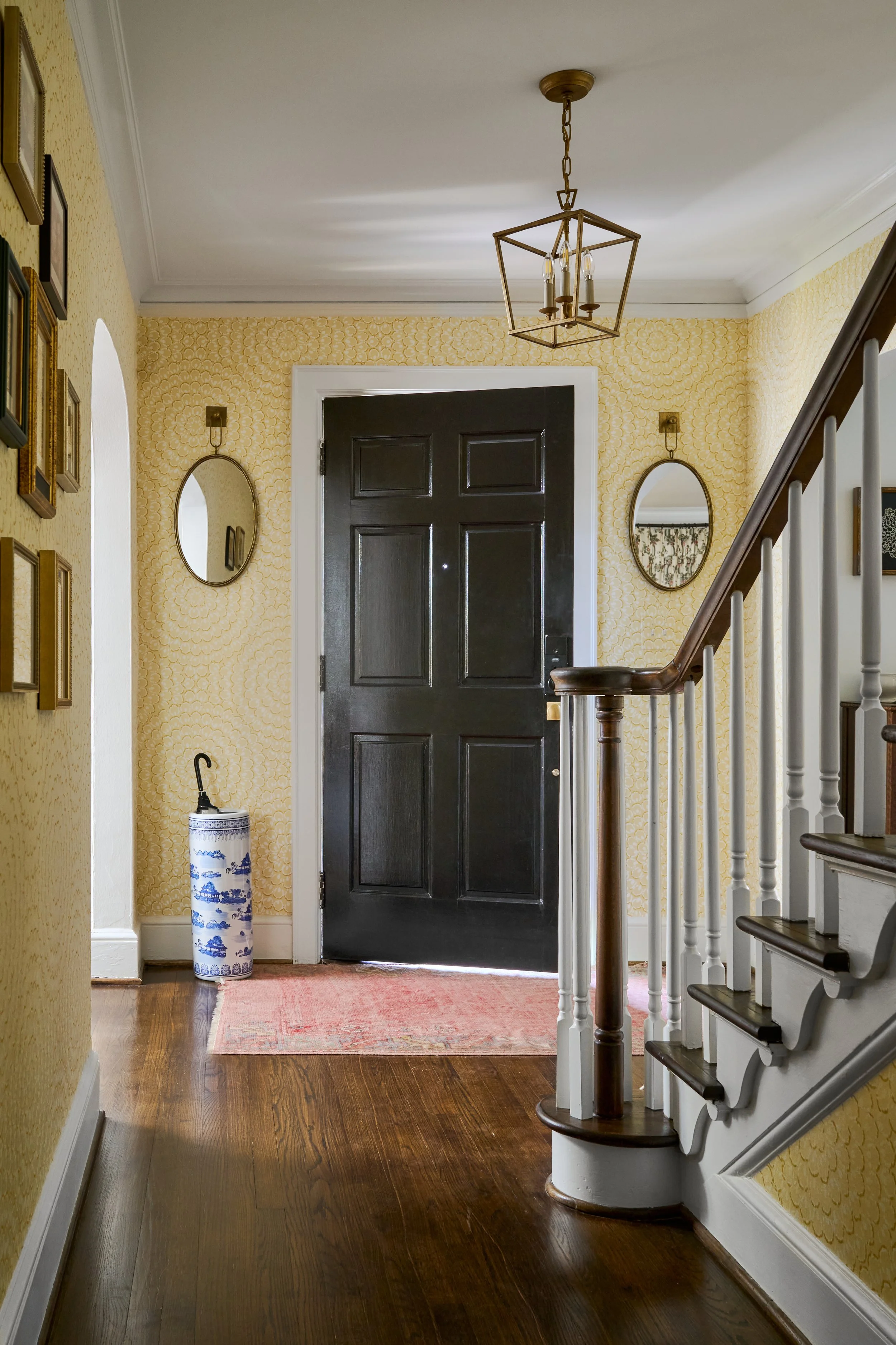 Entryway with black front door, yellow patterned Thibaut wallpaper, oval decorative mirrors sconces, staircase with wooden handrail, hardwood floor, pink rug, and chinoiserie umbrella stand.