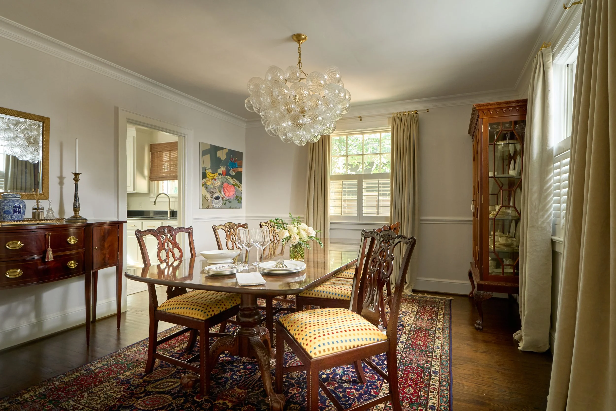 A dining room with a wooden dining table set with plates, glasses, and floral centerpiece, surrounded by upholstered chairs, a chandelier, and wooden cabinet with glass doors, beige curtains, and a patterned rug.