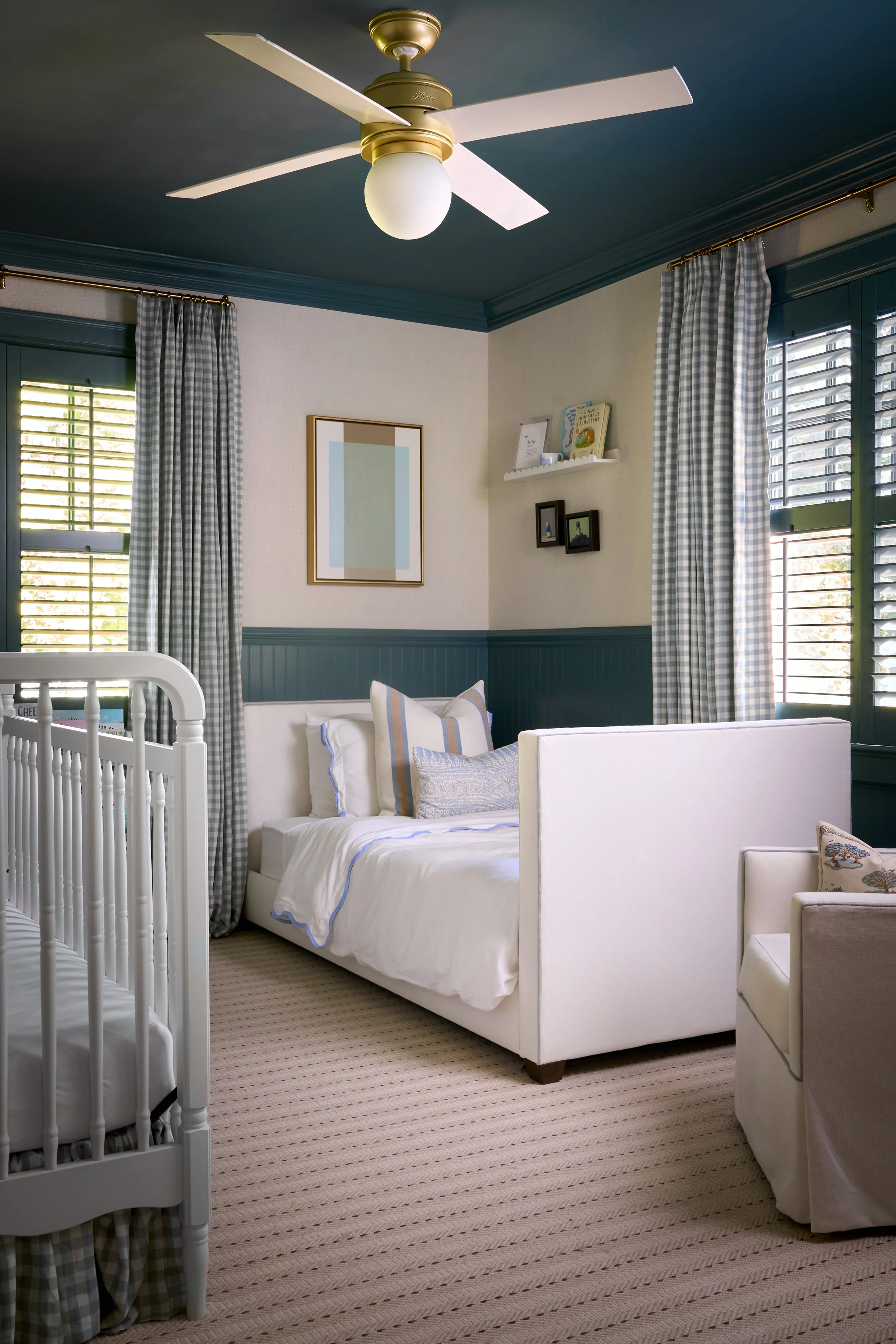 A cozy bedroom with a white bed, striped pillows, and a crib on the left. The room has blue and white checkered curtains, a beige striped rug, and a ceiling fan with a light. There are framed pictures and a shelf with books on the wall.