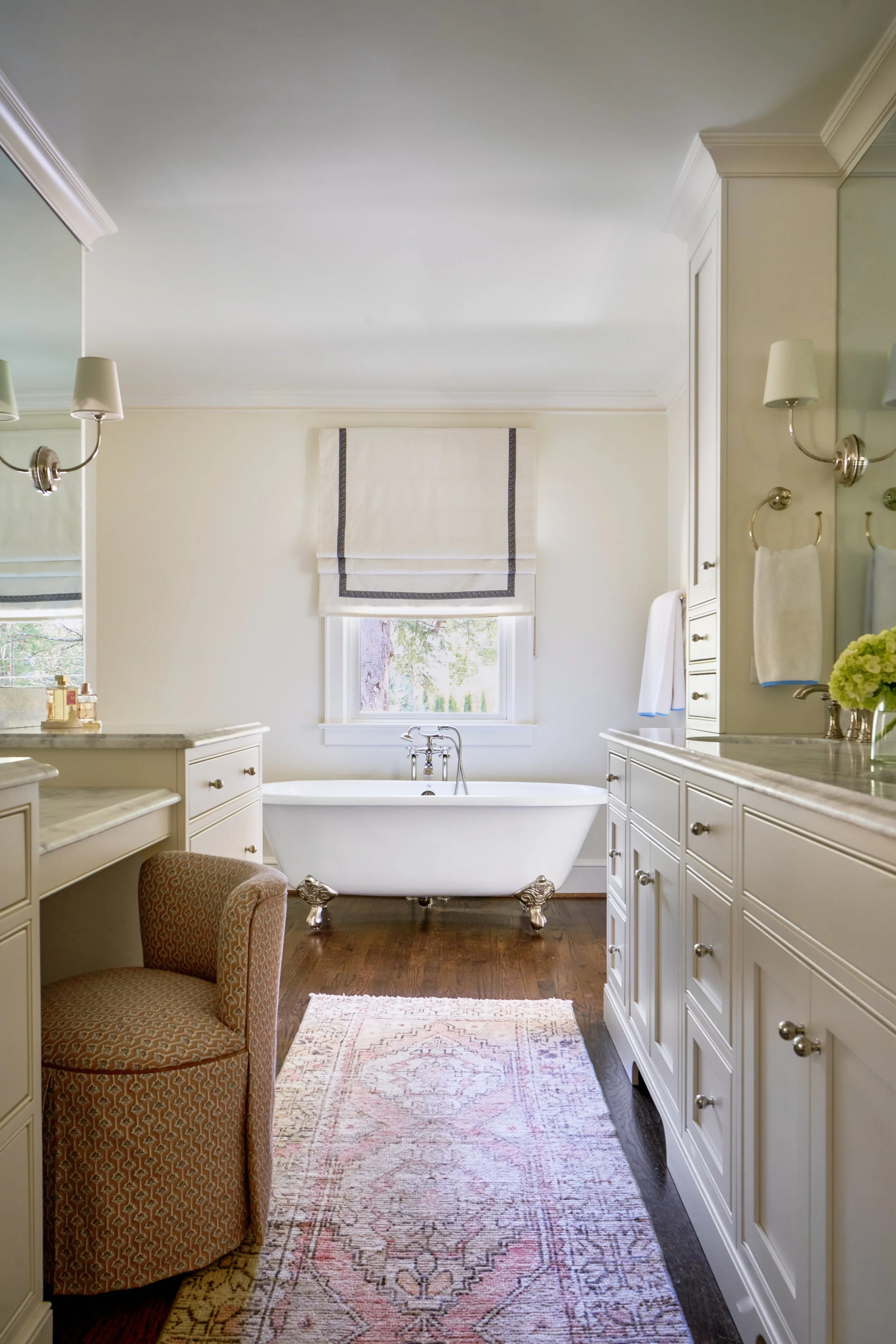 A bathroom featuring a vintage white bathtub with clawfoot design and silver feet, situated below a window with a roman shade. The bathroom has white cabinetry, gold and silver fixtures, and a patterned area rug on dark wood flooring. There are wall-