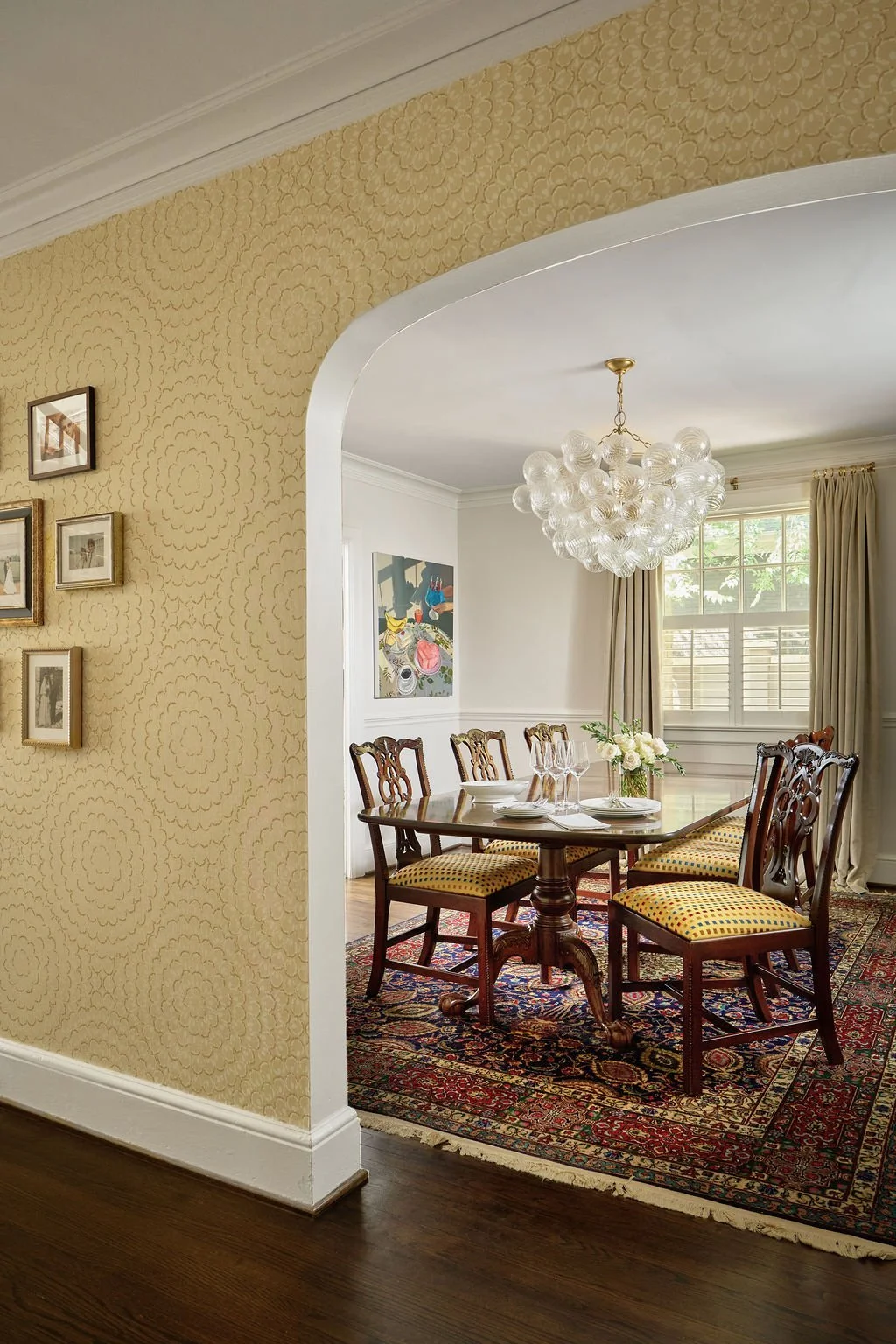 A dining room with Henredon flame mahogany dining table. There are six Chippendale style dining chairs with yellow cushioned seats. The room has a large vintage area rug and beautiful oversized chandelier with multiple glass globes hanging above.