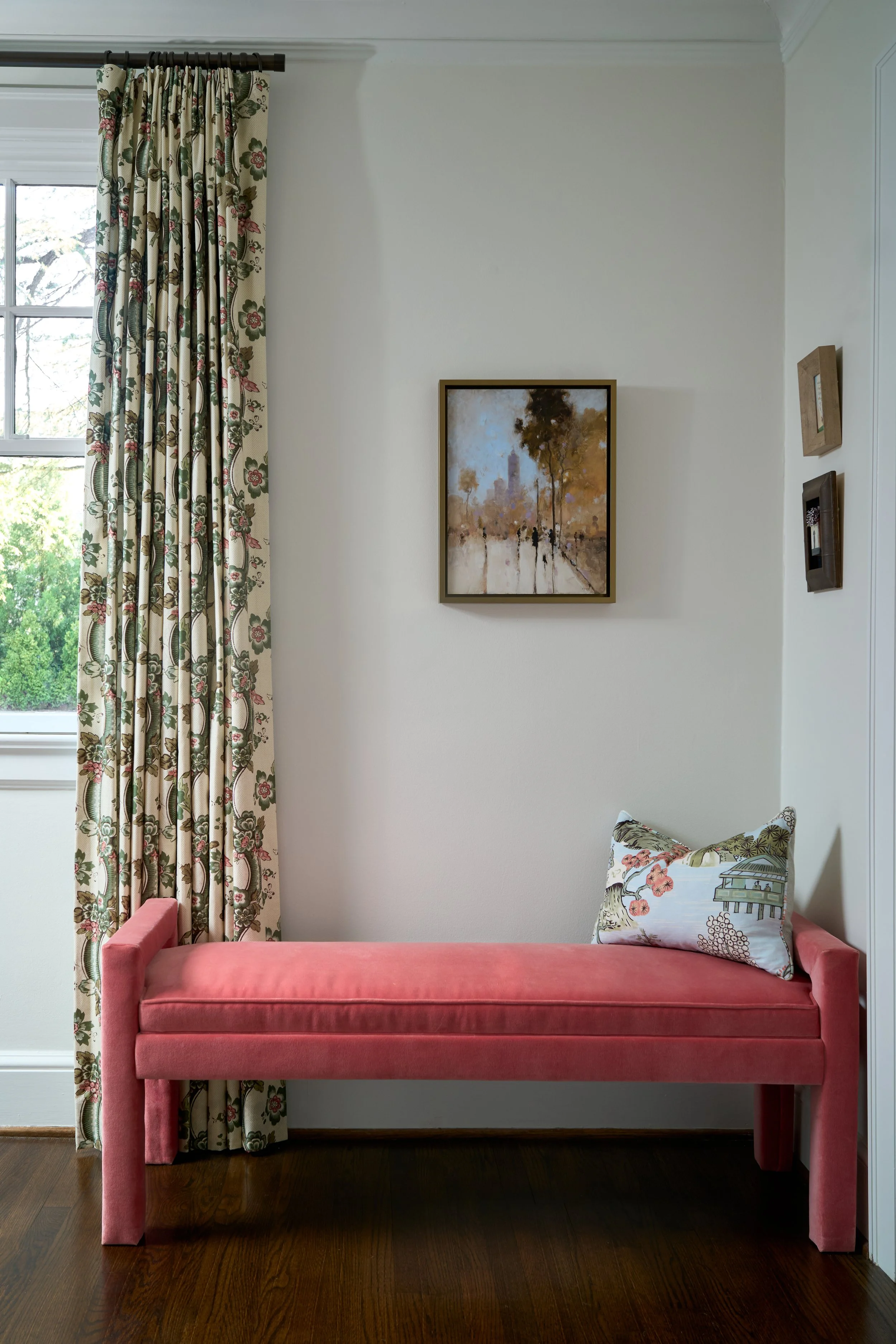 A pink upholstered bench with a decorative pillow featuring a floral and outdoor scene design, placed against a white wall with framed artwork. A window with floral-patterned curtains is on the left side of the image.